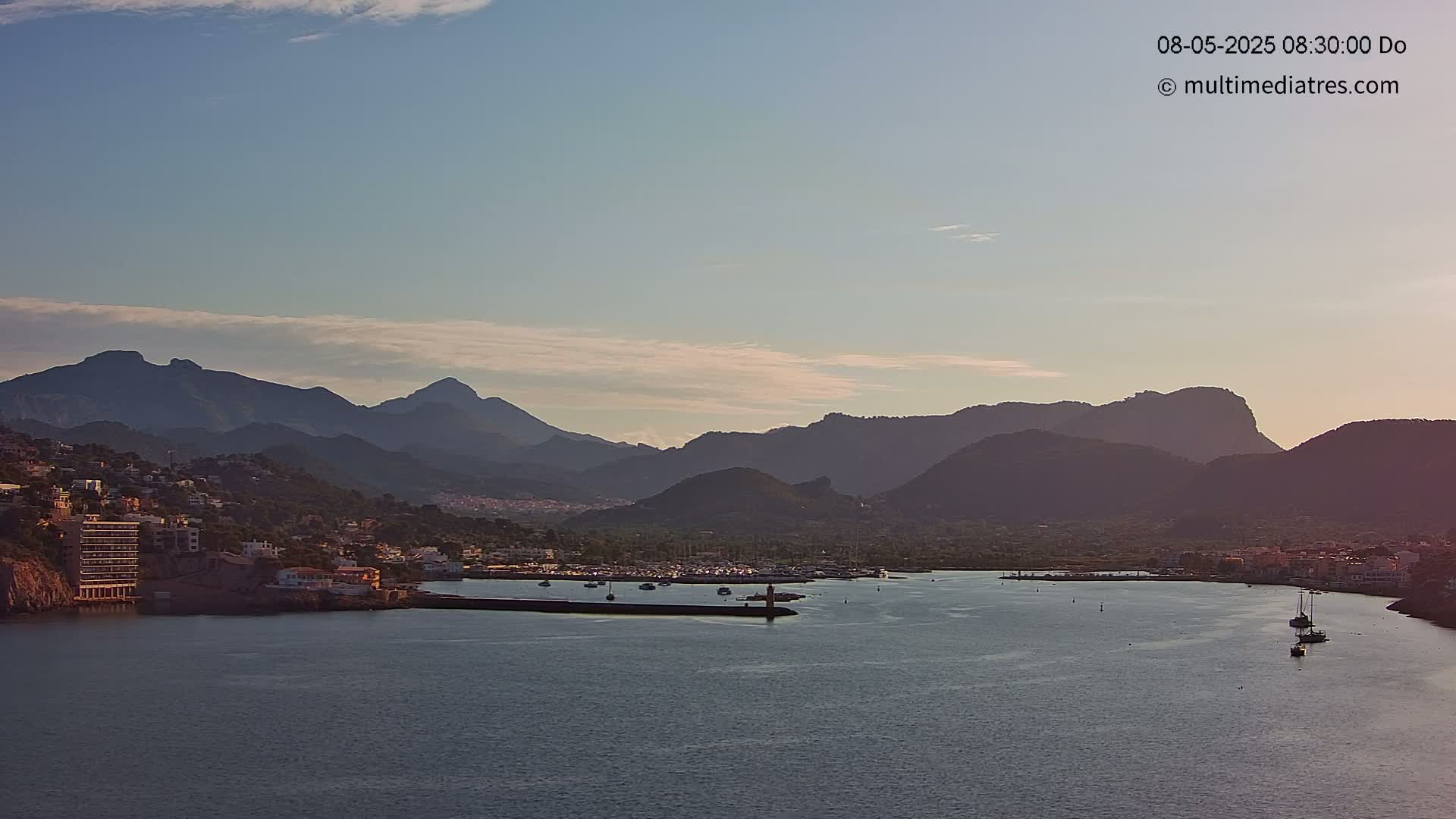 A calm bay with a few boats, a small town nestled along a shoreline, and a mountain range in the background under a mostly clear sky.