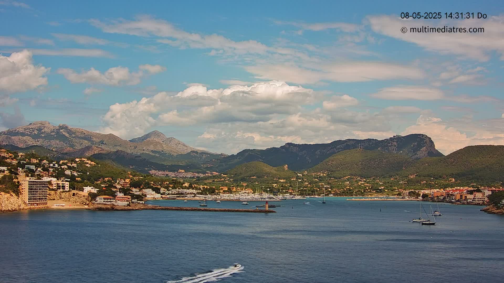 A partly cloudy day reveals a coastal town nestled between a calm bay and verdant mountains.