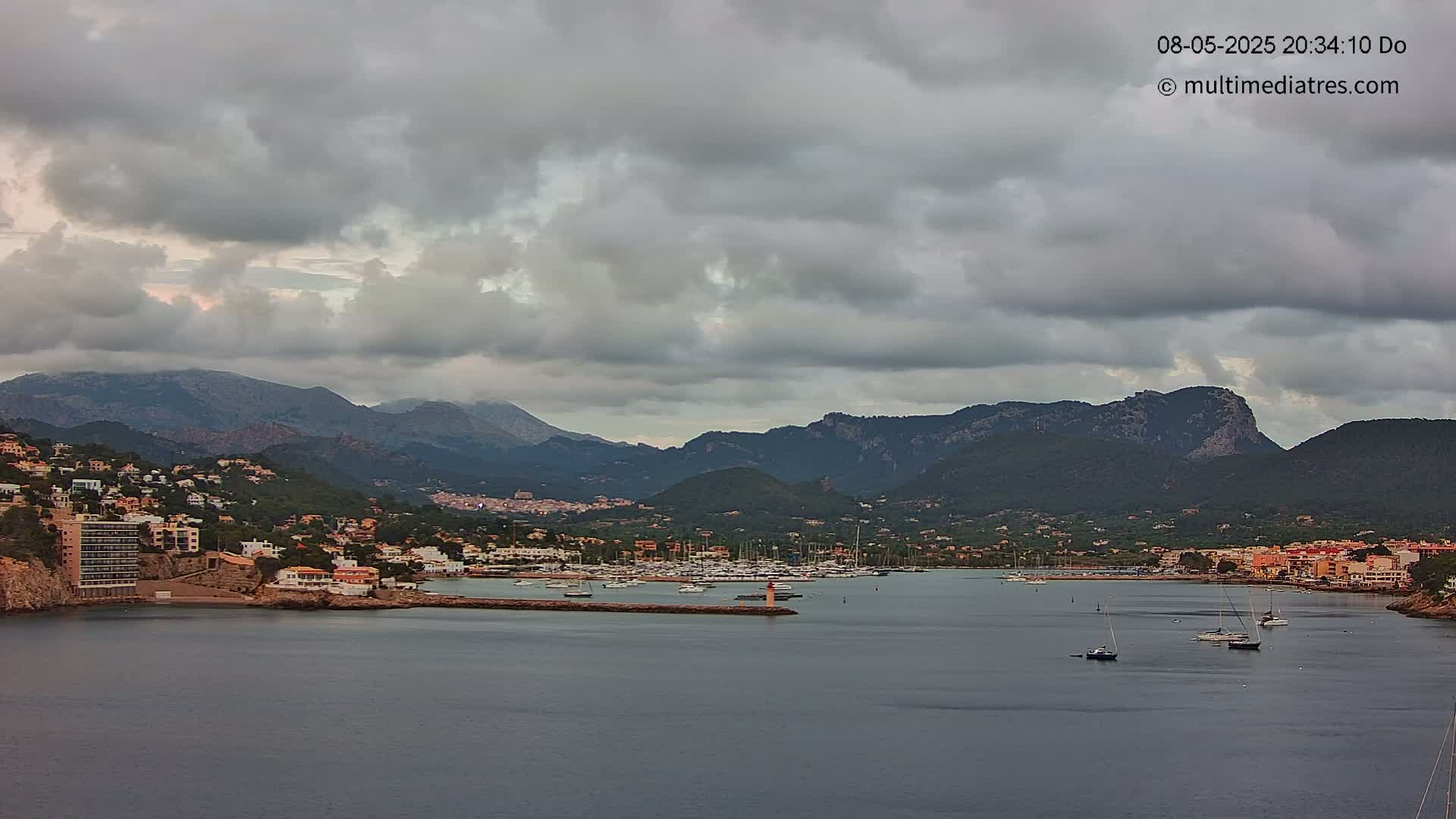 A coastal town nestled in mountains under a cloudy sky overlooks a calm bay with several sailboats.