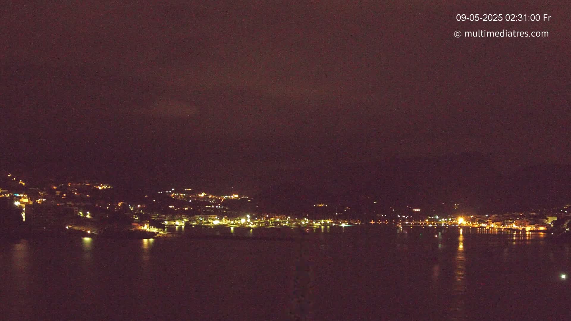 A dark, nighttime view of a town's lights across a body of water under a mostly clear sky.