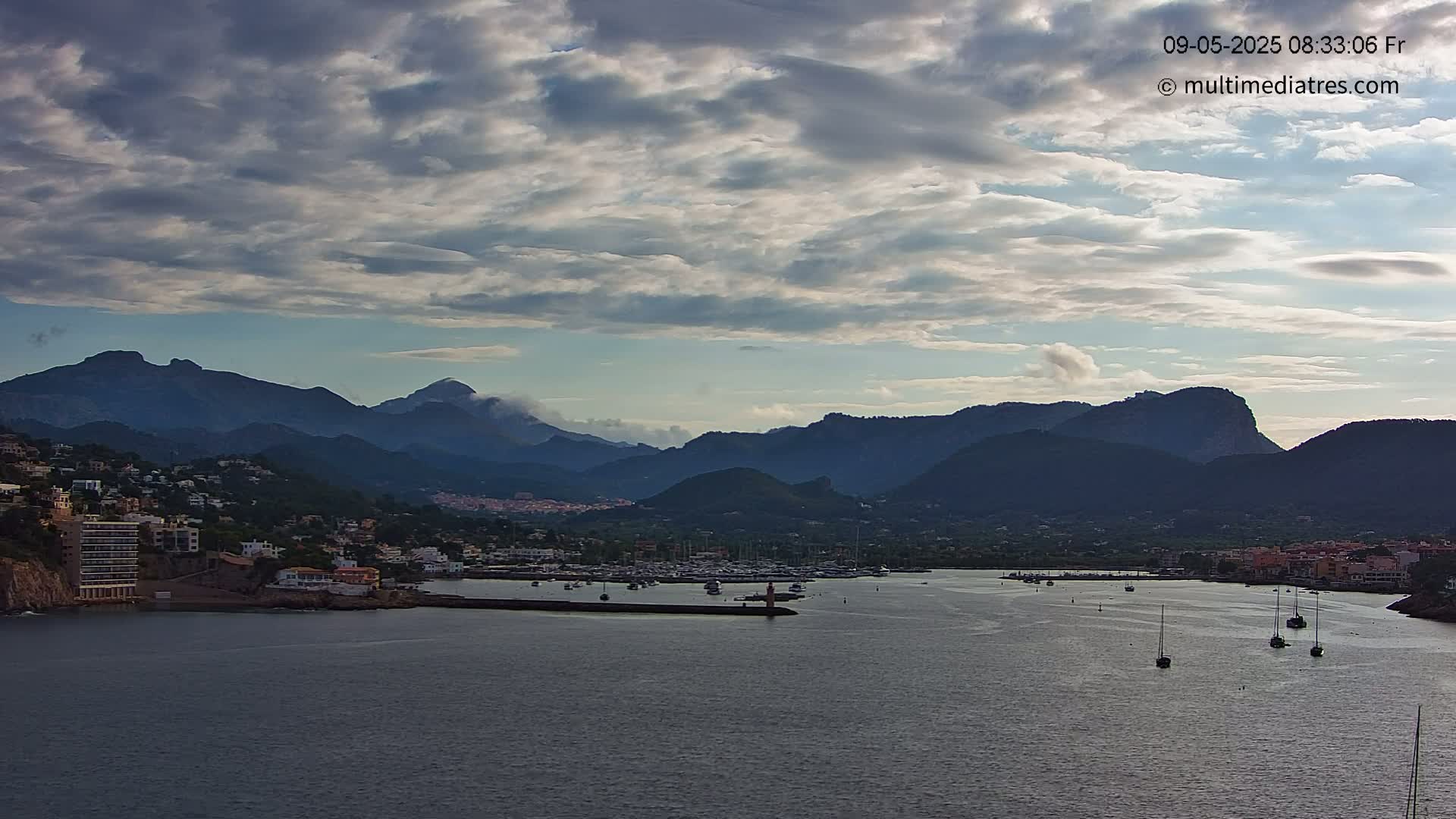 A partly cloudy sky overlooks a harbor filled with boats, nestled between a town and a mountain range.