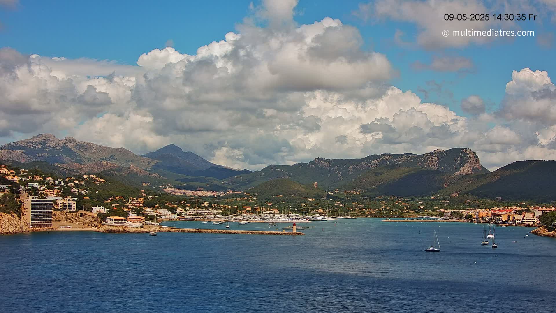 A partly cloudy sky overlooks a coastal town nestled against a mountain range, with a calm bay containing several sailboats.