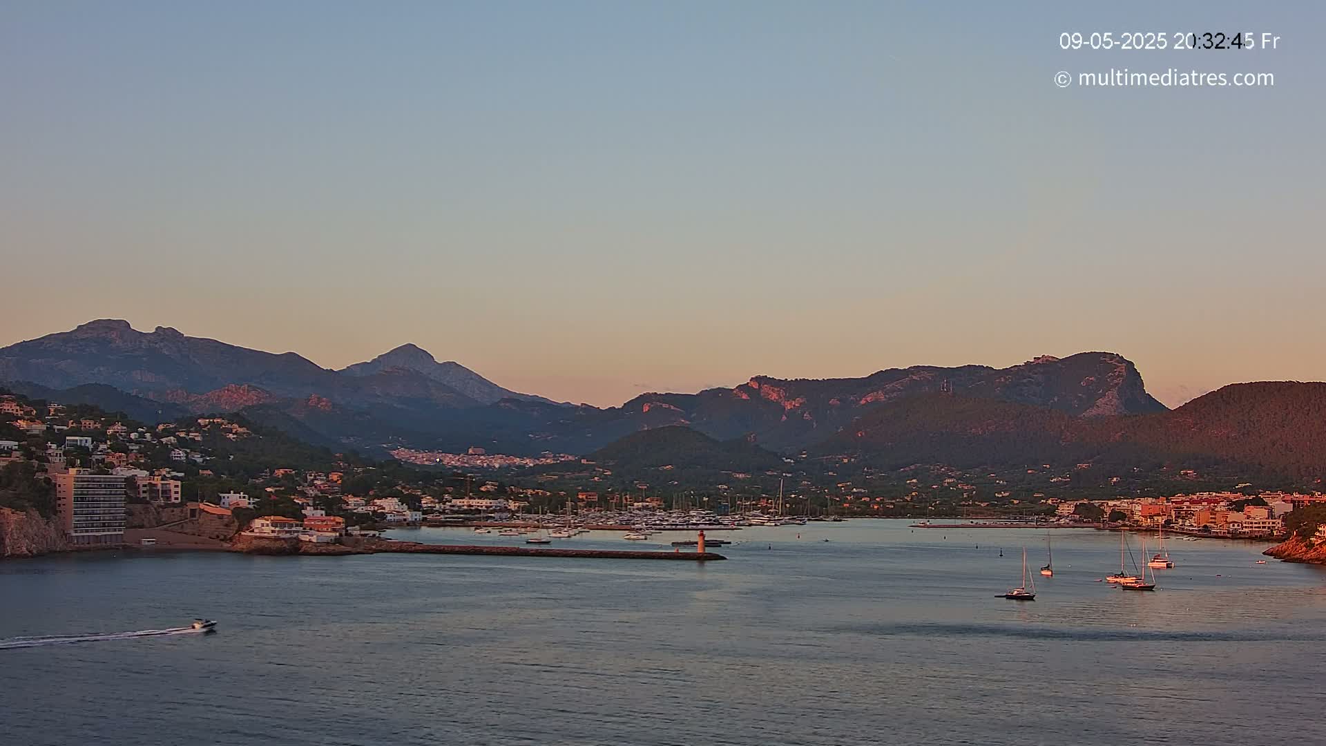 A coastal town nestled at the foot of mountains is bathed in the soft light of sunset, with several boats anchored in the calm water of a harbor.