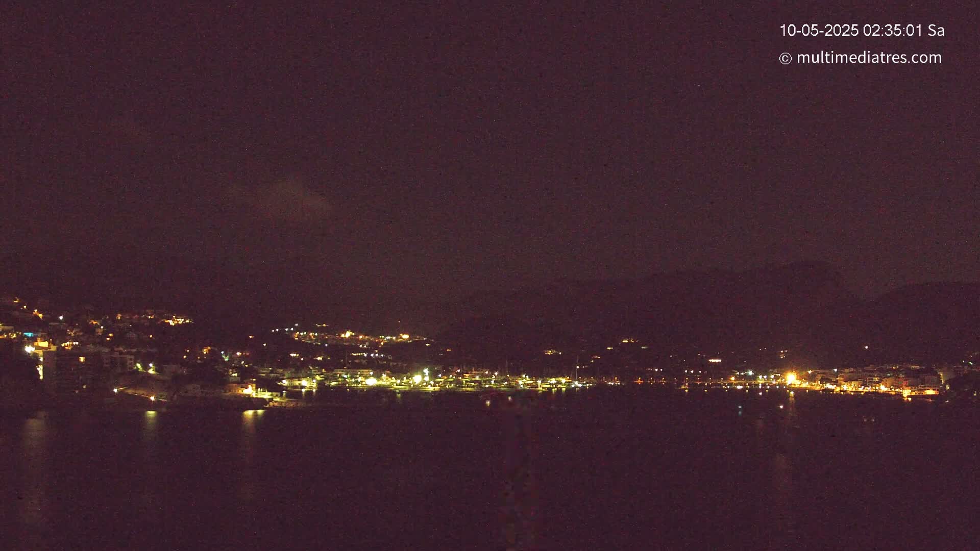 A nighttime view of a coastal town with many lights visible across a dark body of water under a clear, dark sky.
