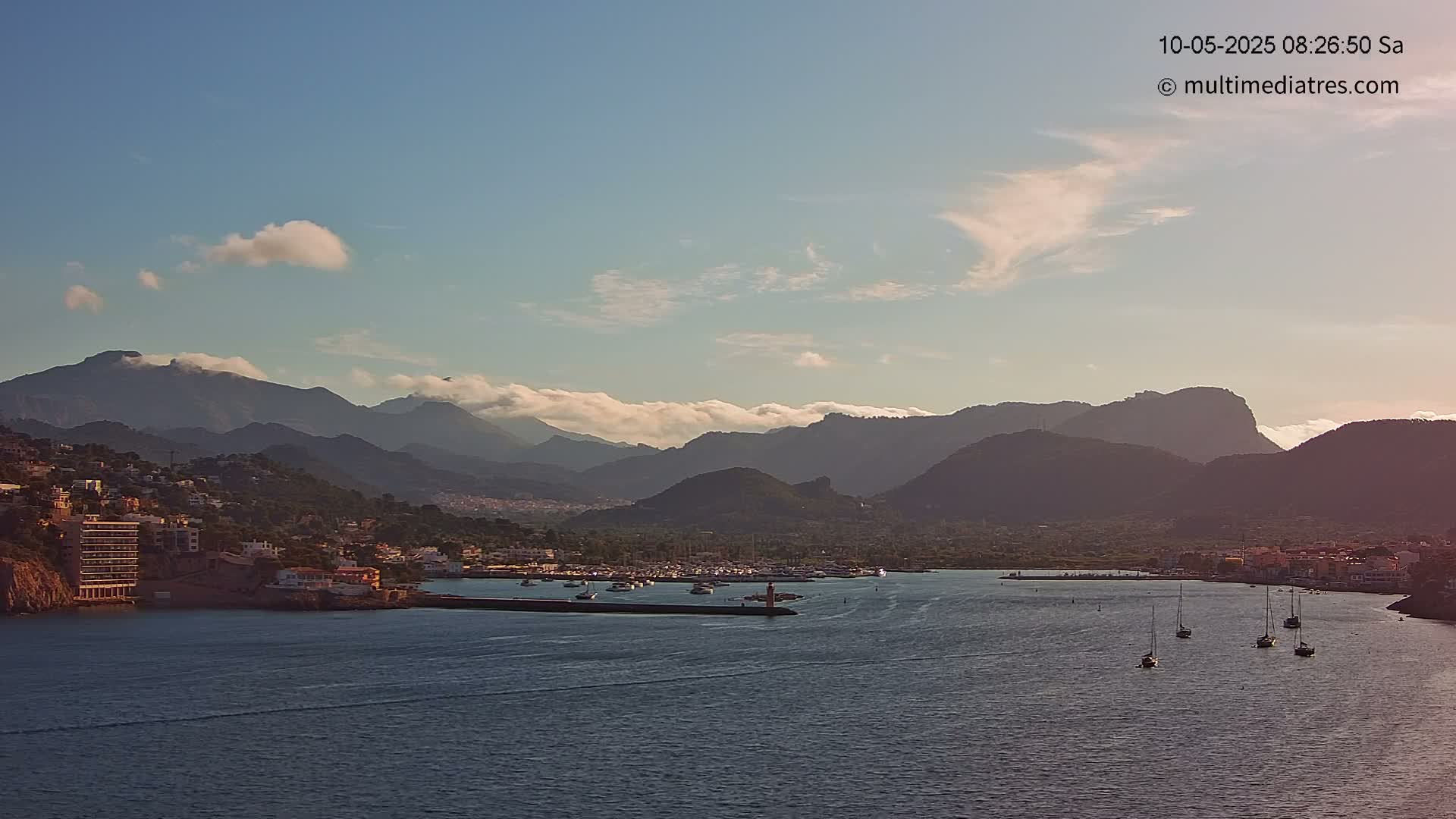 A calm body of water with several sailboats is nestled in a harbor next to a town, which sits at the base of a mountain range partially covered in clouds under a clear sky.
