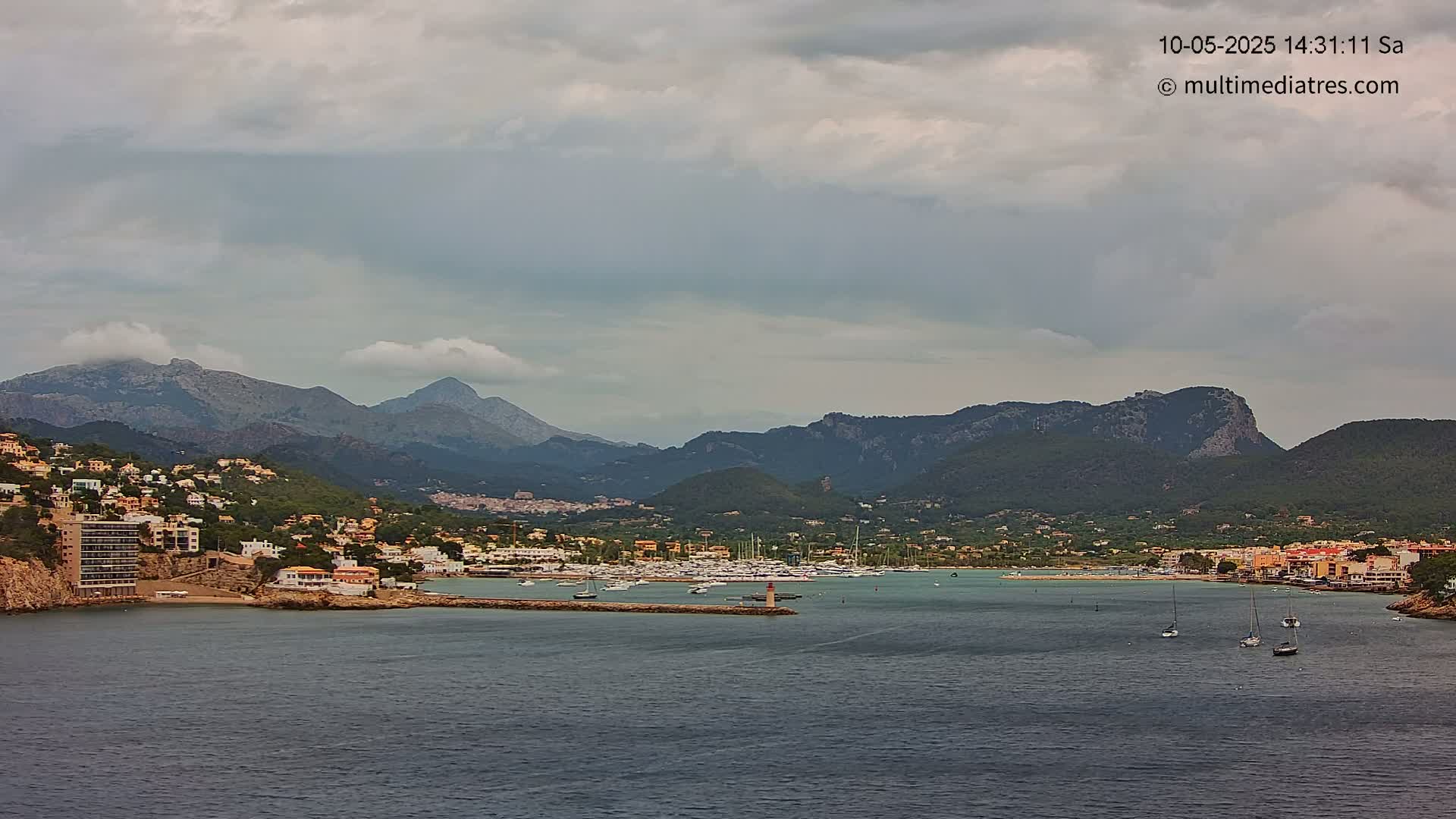 A coastal town nestled amongst mountains under a cloudy sky, with a harbor filled with boats.