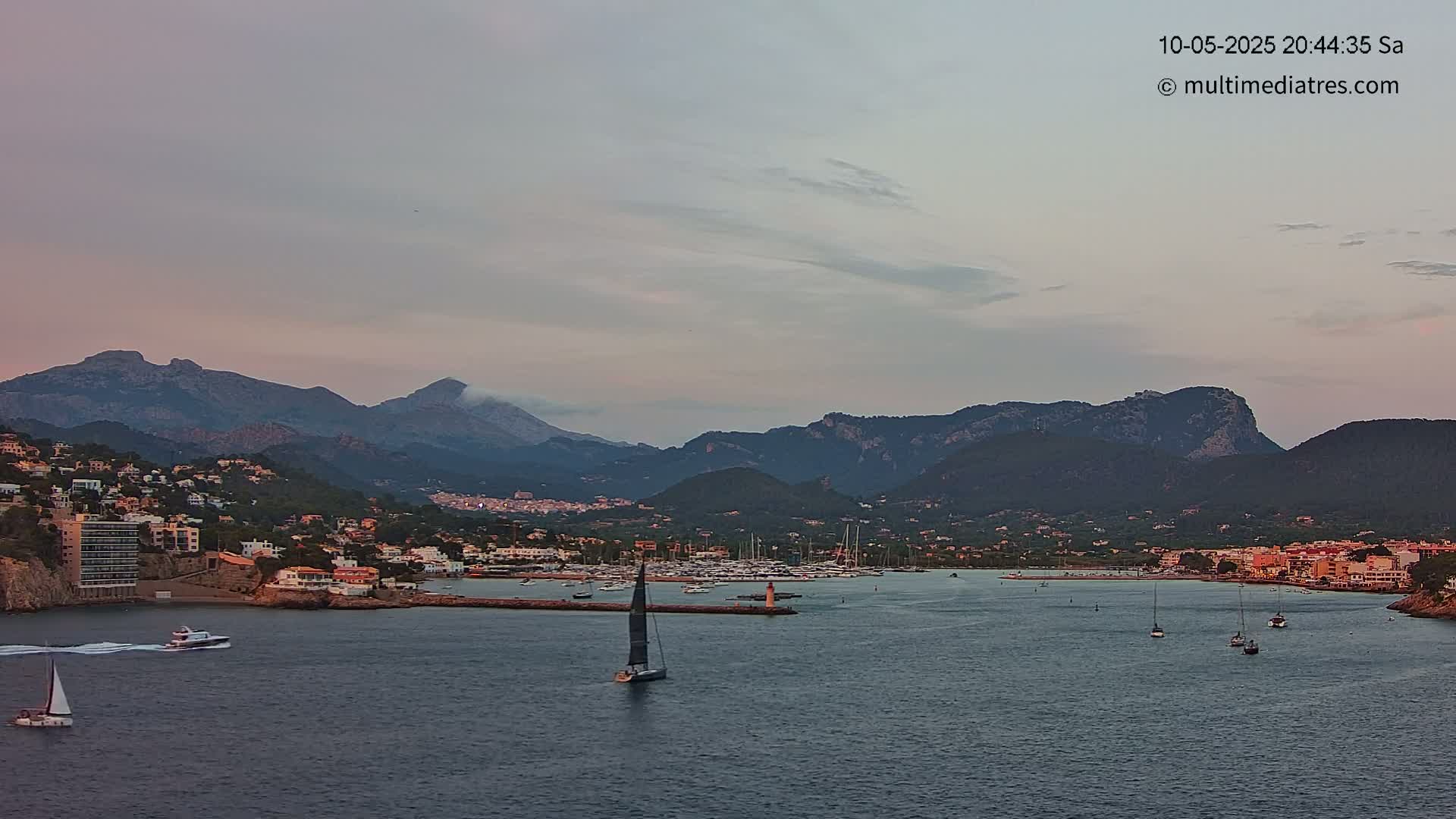 A calm, twilight-lit harbor is nestled between coastal buildings and a range of dark mountains under a pale sky.