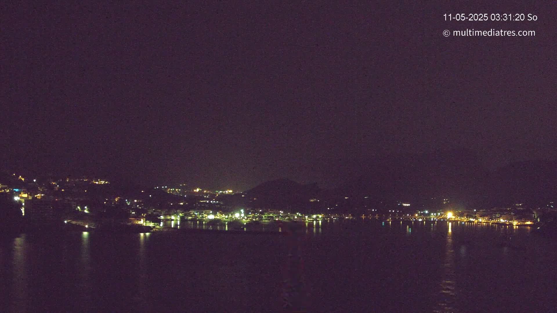 A nighttime view of a town's lights spread across a hillside, reflected in a dark body of water under a clear, dark sky.
