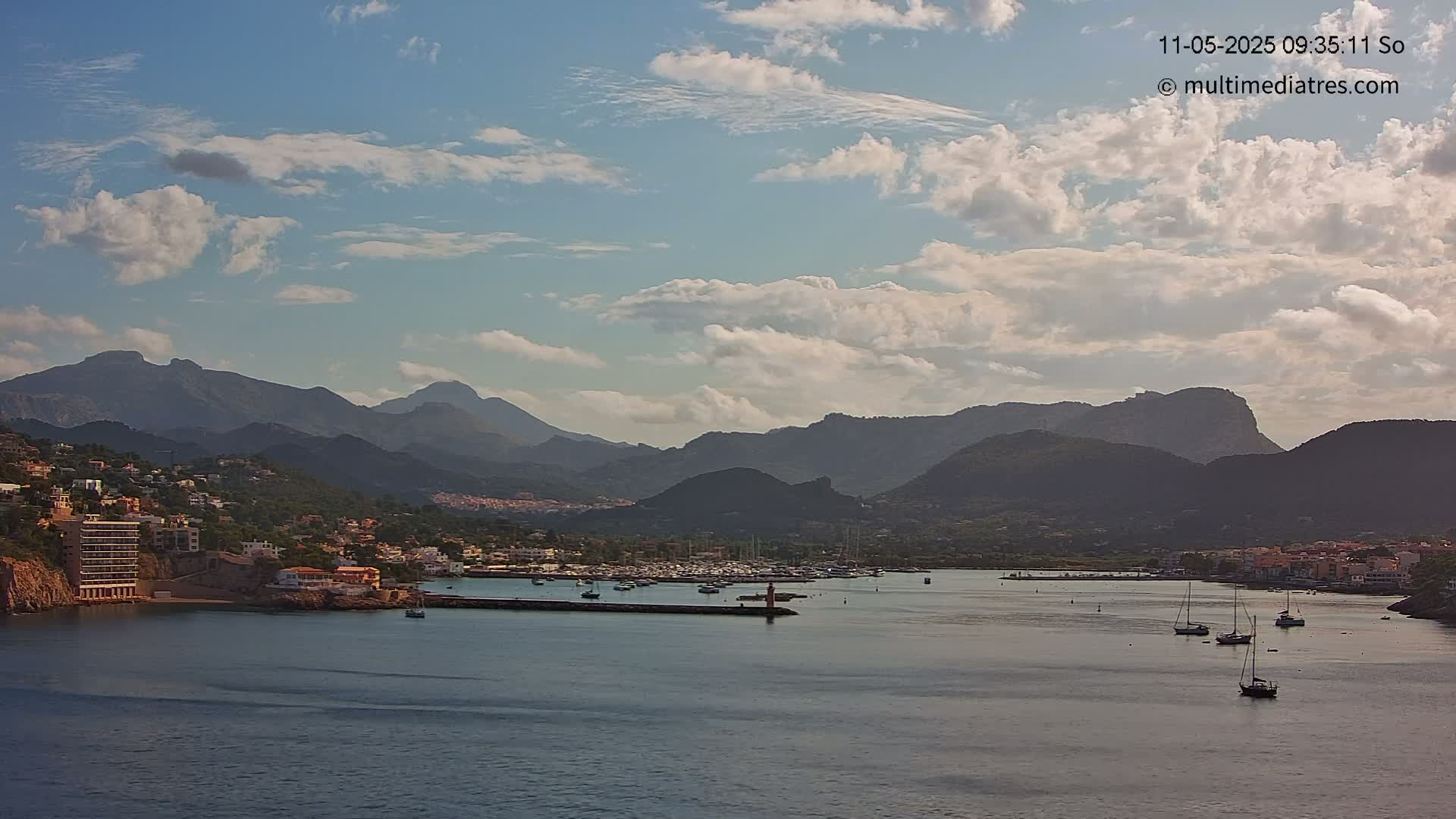 A partly cloudy sky overlooks a calm bay with numerous boats, a small town nestled on the shore, and mountains in the background.