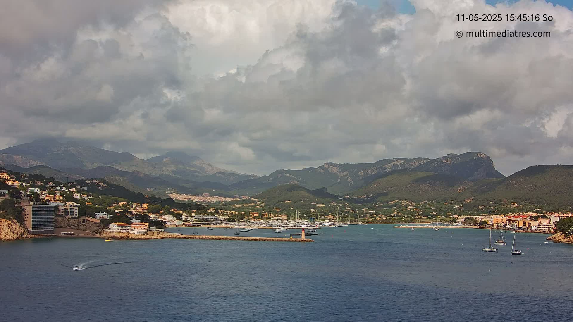 A coastal town nestled against a mountainous backdrop under a cloudy sky, with a harbor filled with boats.