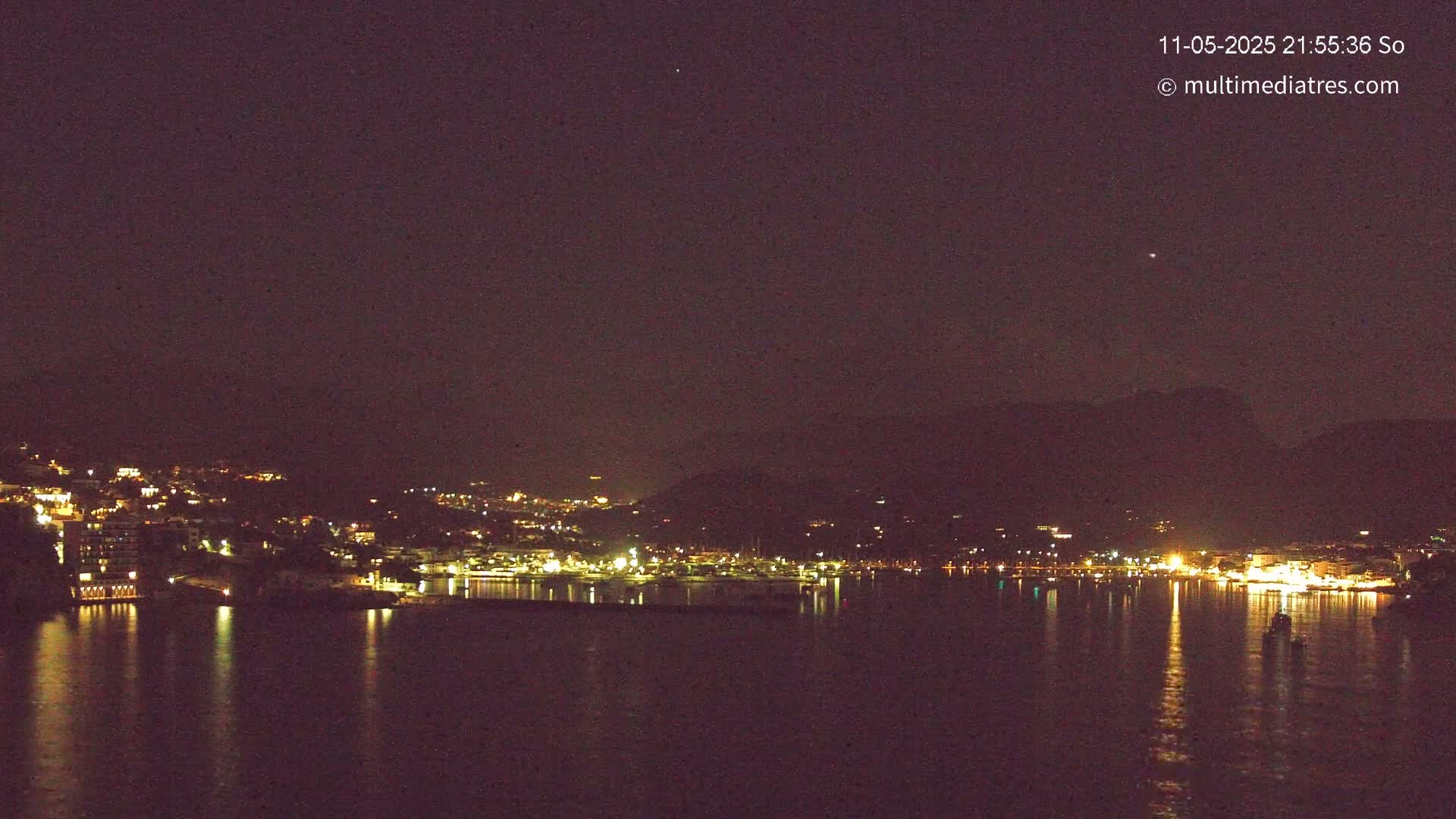 A nighttime view of a town's lights illuminating a dark body of water, with hills in the background under a clear, dark sky.