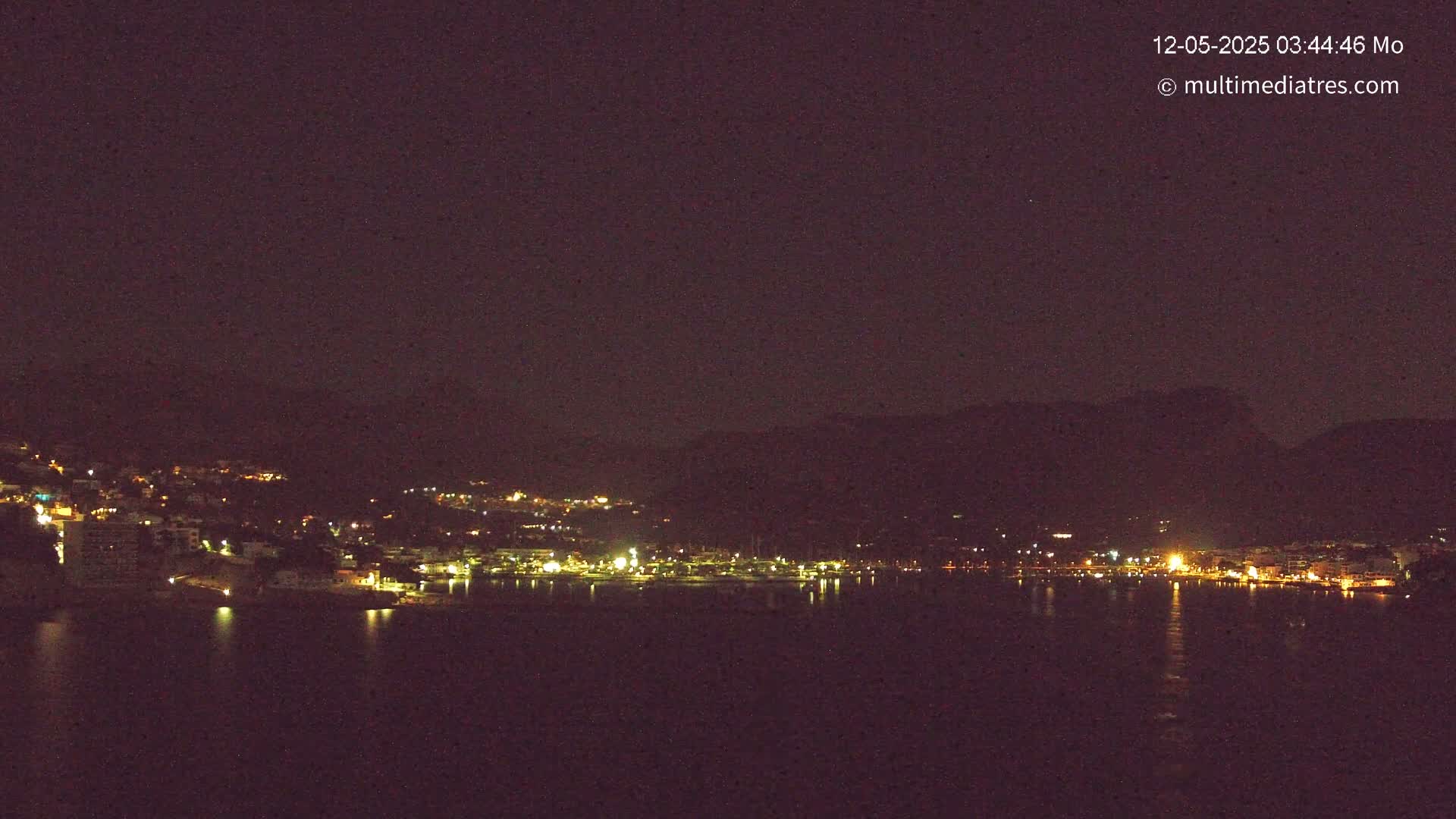A dark, nighttime view of a coastal town illuminated by scattered lights, nestled against a dark mountain range, across a calm body of water.