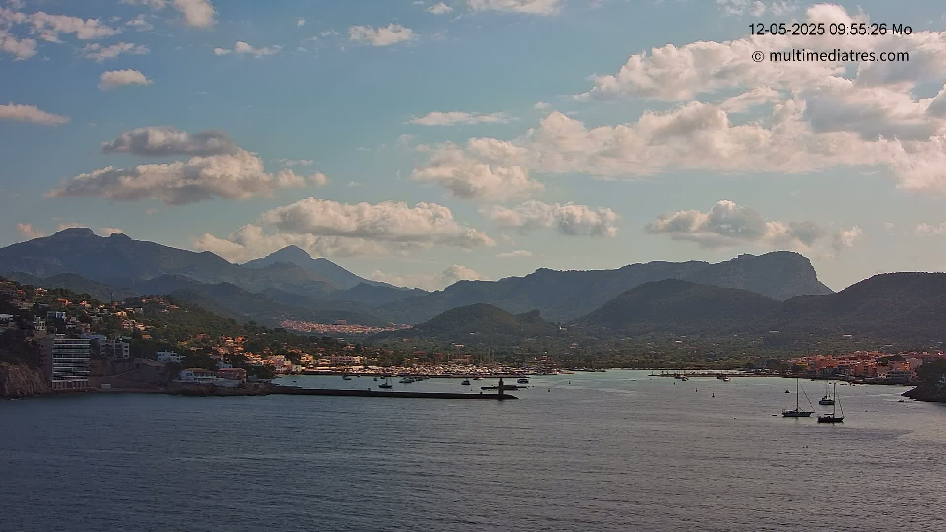 A partly cloudy day shows a coastal town nestled between mountains and a calm bay with several boats.