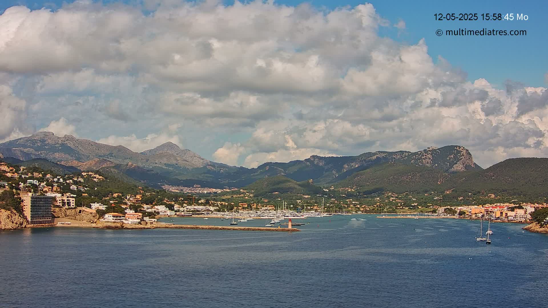 A partly cloudy day reveals a coastal town nestled between mountains and a calm bay with a few sailboats.