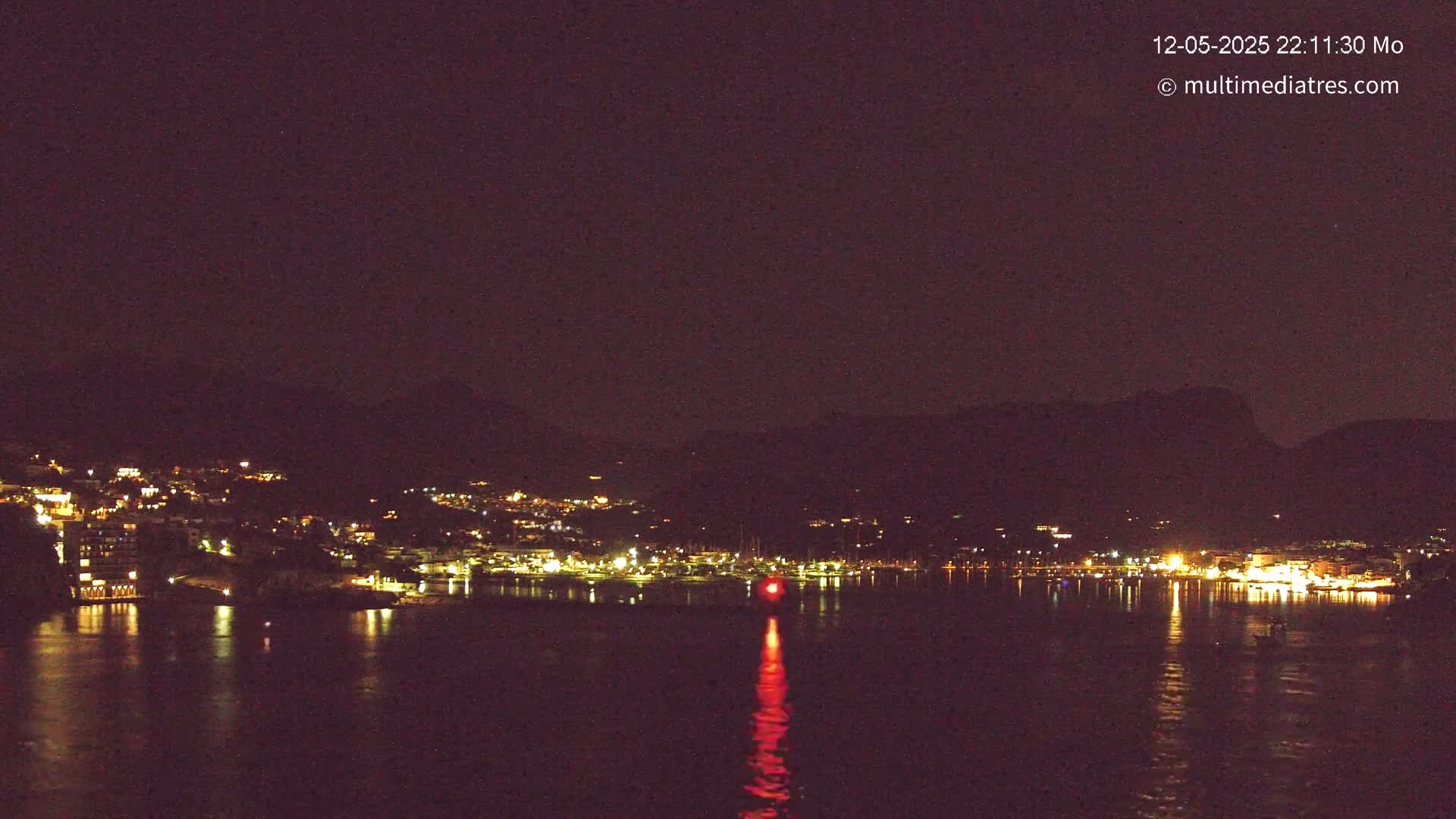 A nighttime view of a city's lights across a dark body of water, nestled between hills under a clear, dark sky.