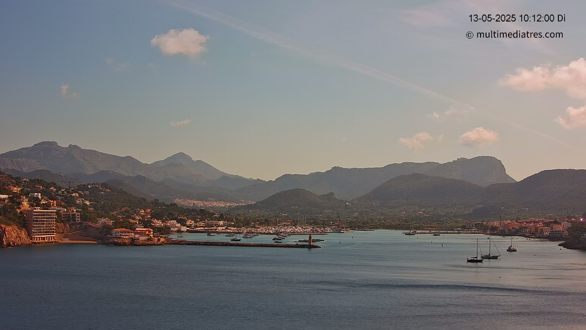 A harbor filled with boats is nestled between a coastal town and a mountain range under a partly cloudy sky.