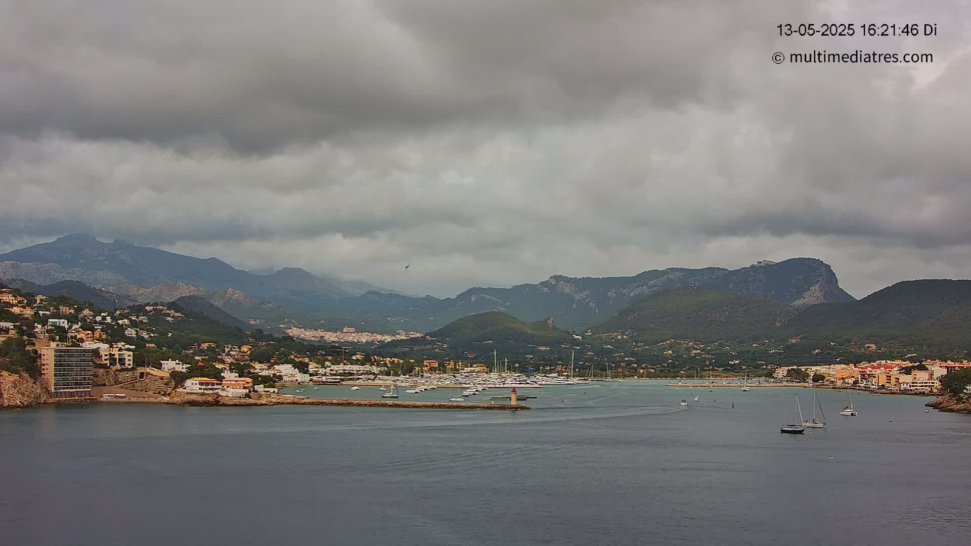A coastal town nestled between mountains under a cloudy sky, with a harbor filled with boats.