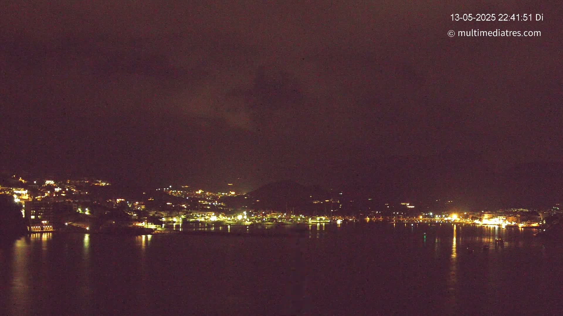 A nighttime view of a town on a hillside, with its lights reflected on a calm body of water under a dark, cloudy sky.