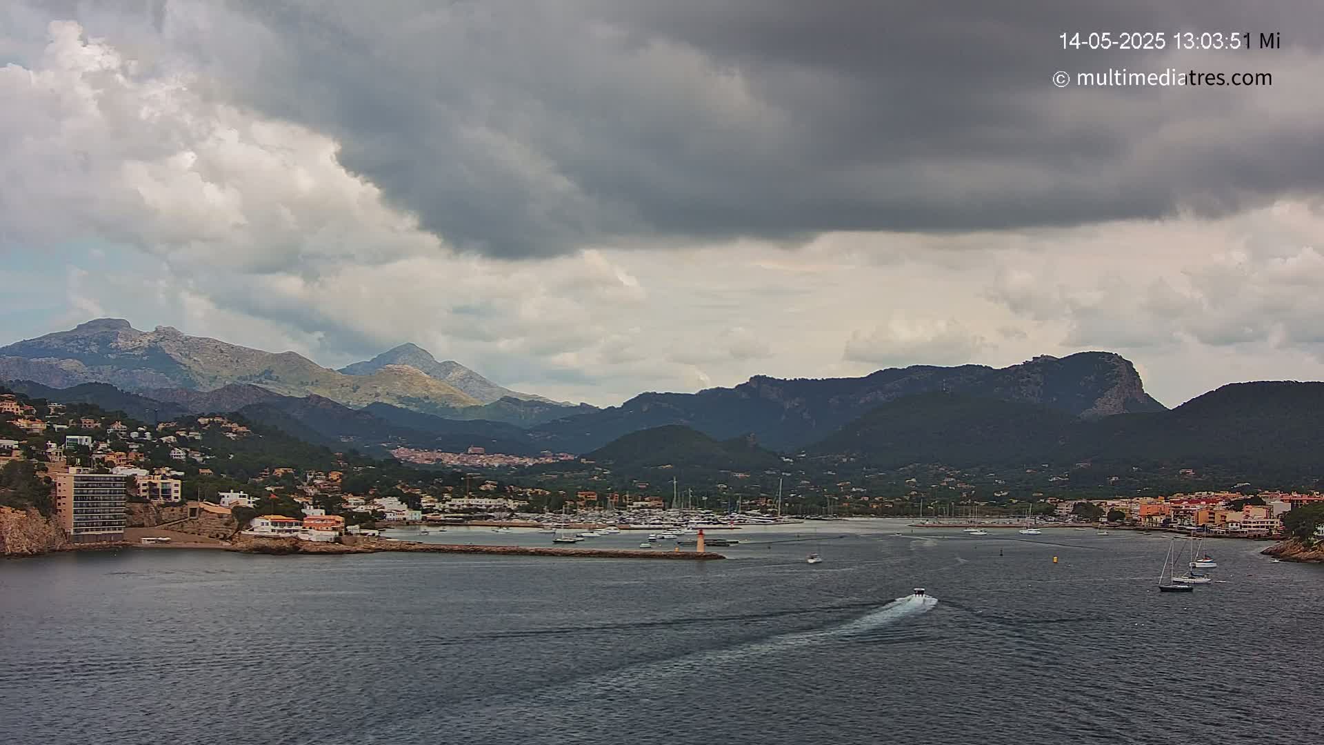 A coastal town nestled between dark, mountainous terrain under a mostly cloudy sky, with a calm body of water featuring several boats.