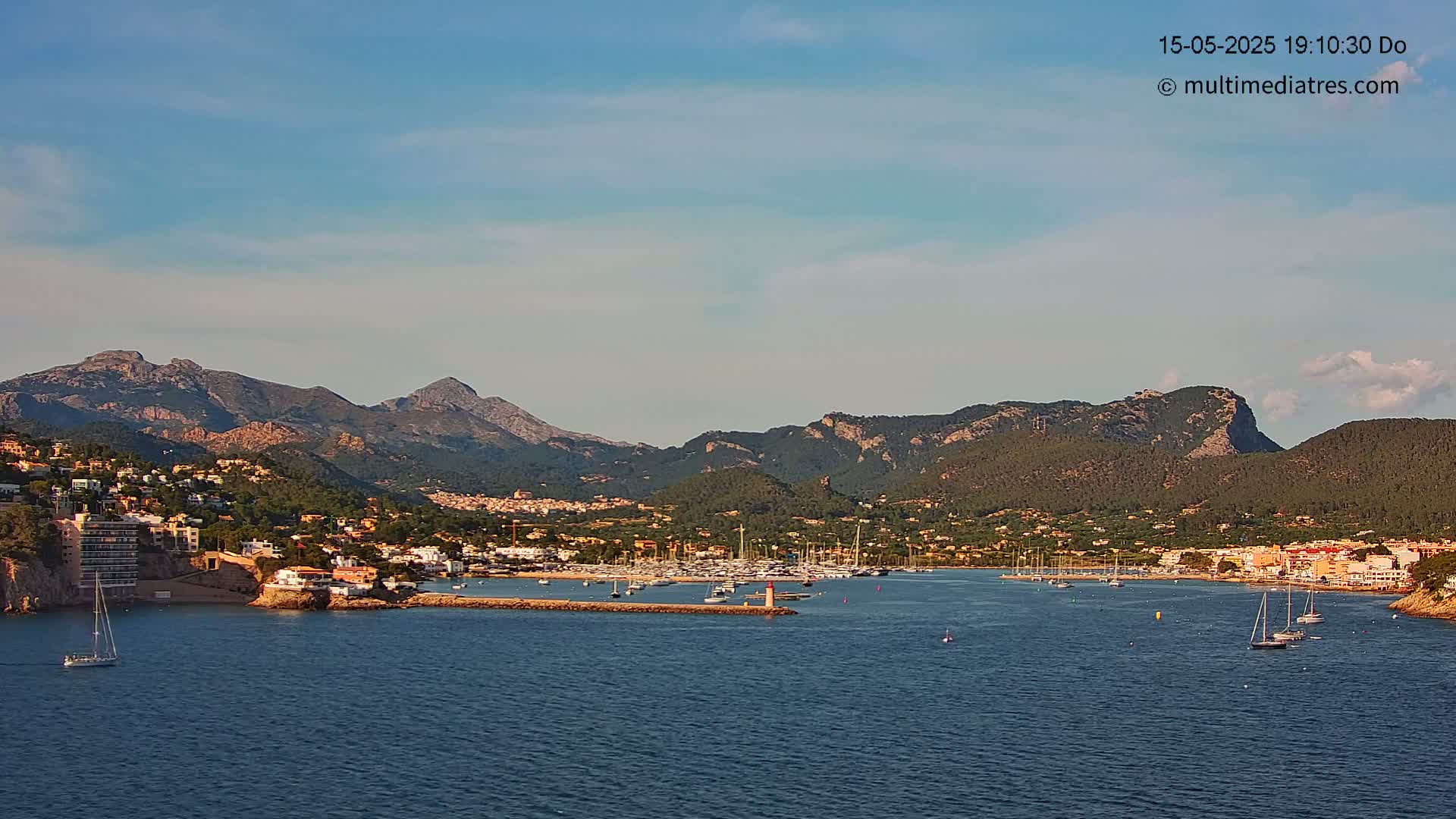 A coastal town nestled between mountains, with a marina filled with sailboats, under a mostly clear sky.