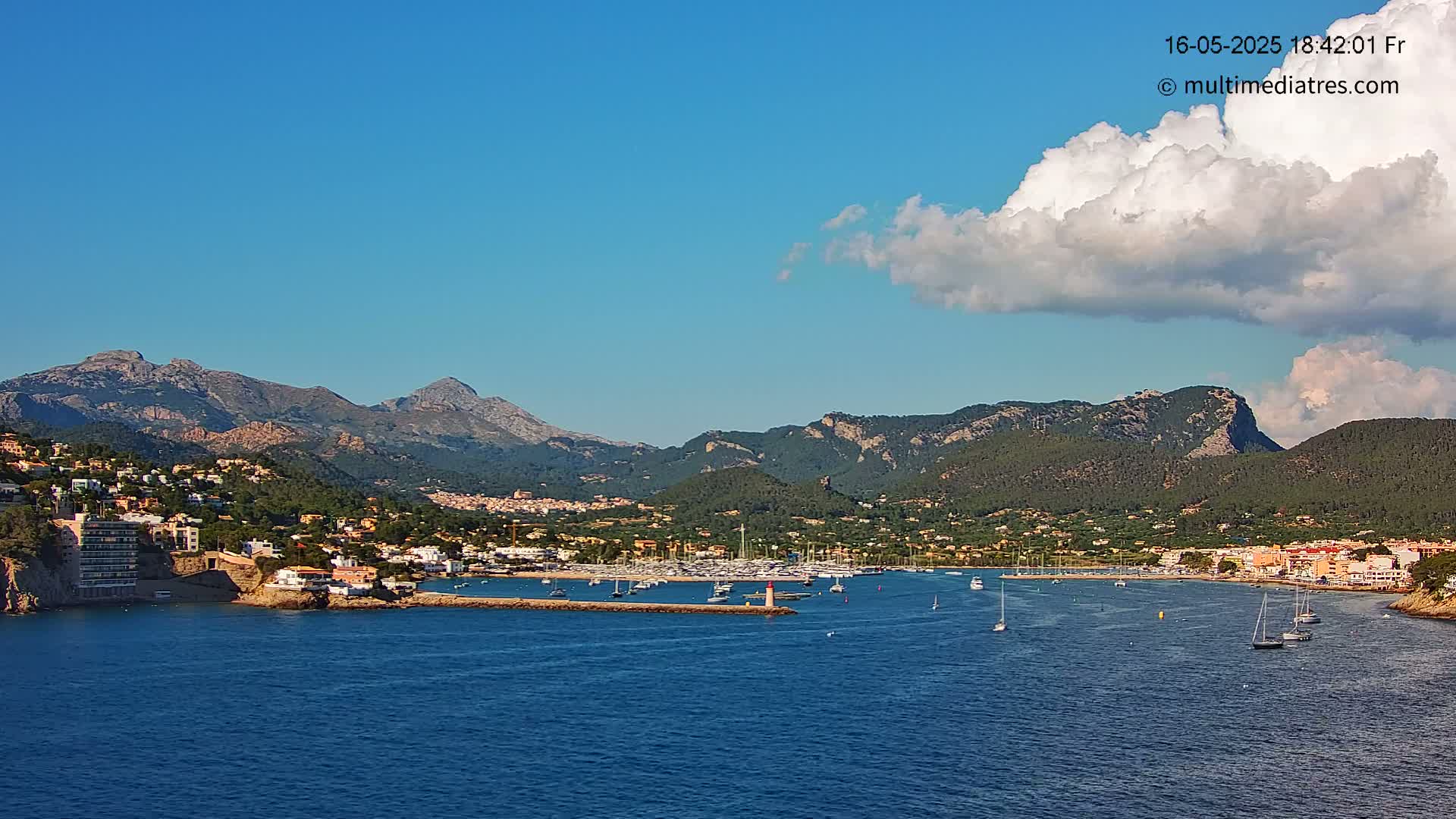 A coastal town nestled against mountainous terrain under a mostly clear blue sky with a single large cumulus cloud.