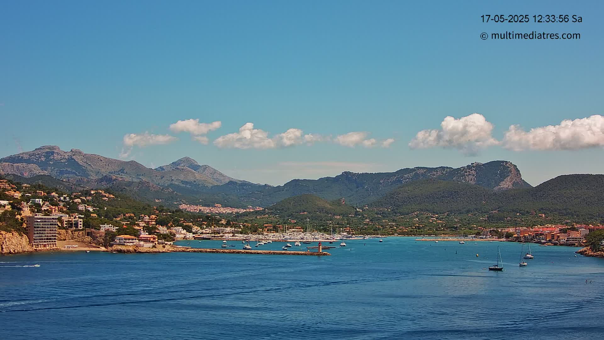 A picturesque coastal town nestled against a backdrop of mountains under a mostly clear, sunny sky with a few scattered clouds, features a calm bay filled with sailboats.