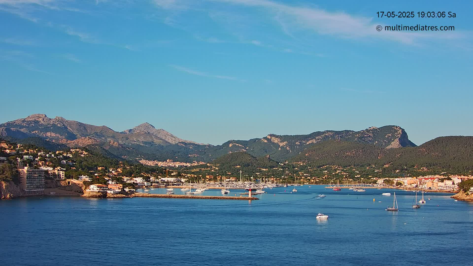 A picturesque harbor filled with boats is nestled between a coastal town and a range of mountains under a clear blue sky.
