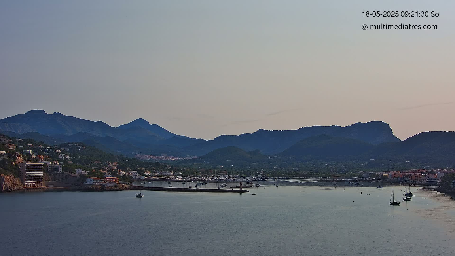 A calm bay with a small marina nestled between a hillside town and a range of dark mountains under a clear, light-grey sky.