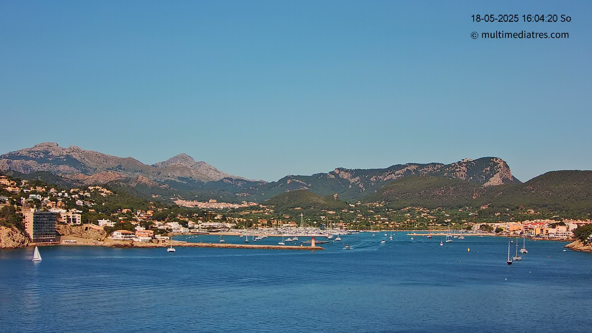 A coastal town nestled between mountains and a calm blue sea under a clear, sunny sky.