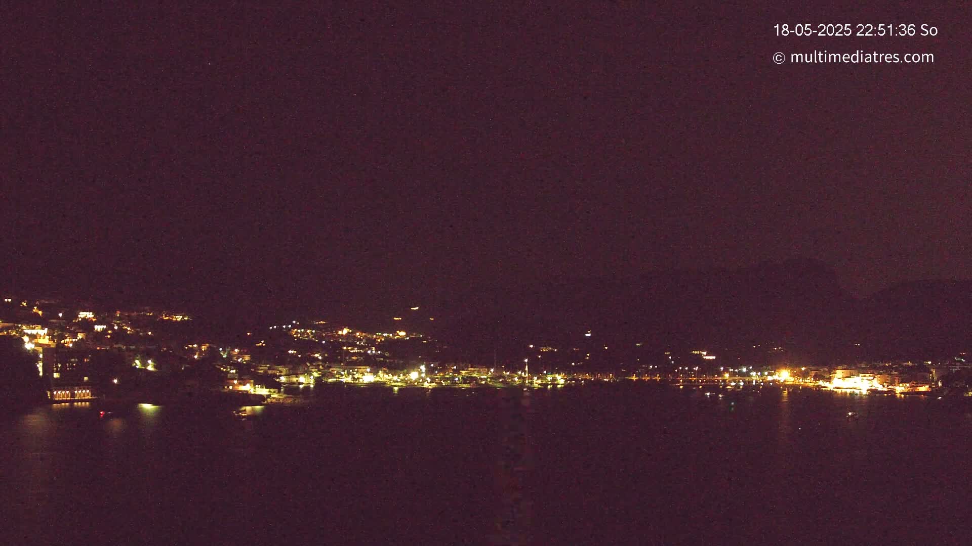A nighttime view of a coastal town illuminated by many lights, seen across a dark body of water under a clear, dark sky.