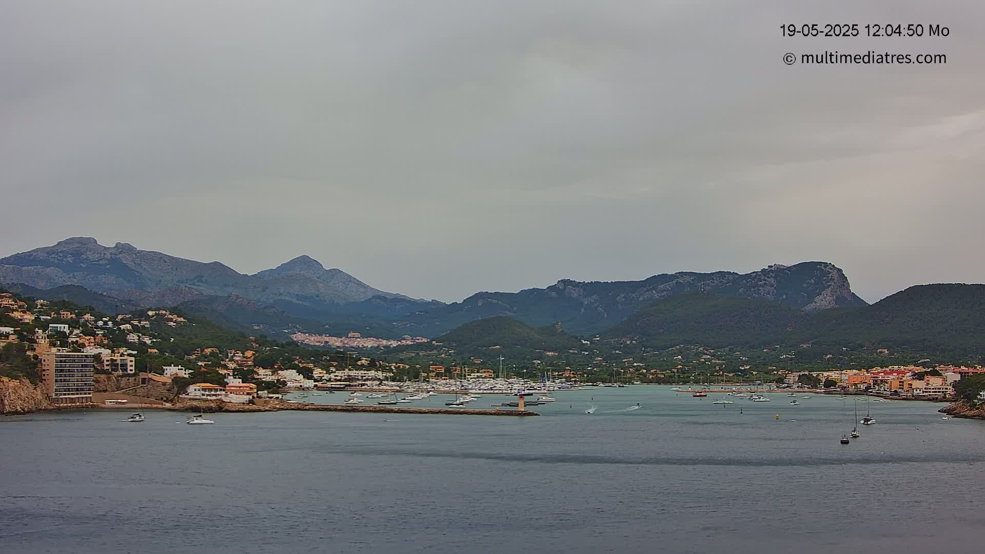 A coastal town nestled at the foot of a mountain range under an overcast sky, with a calm harbor filled with boats.