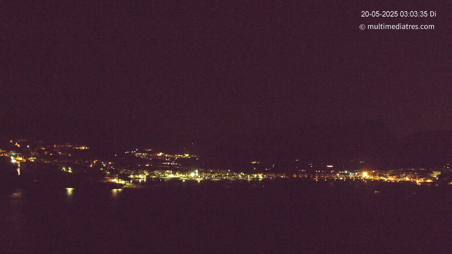 A nighttime view of a coastal town's lights across a dark body of water under a clear, dark sky.