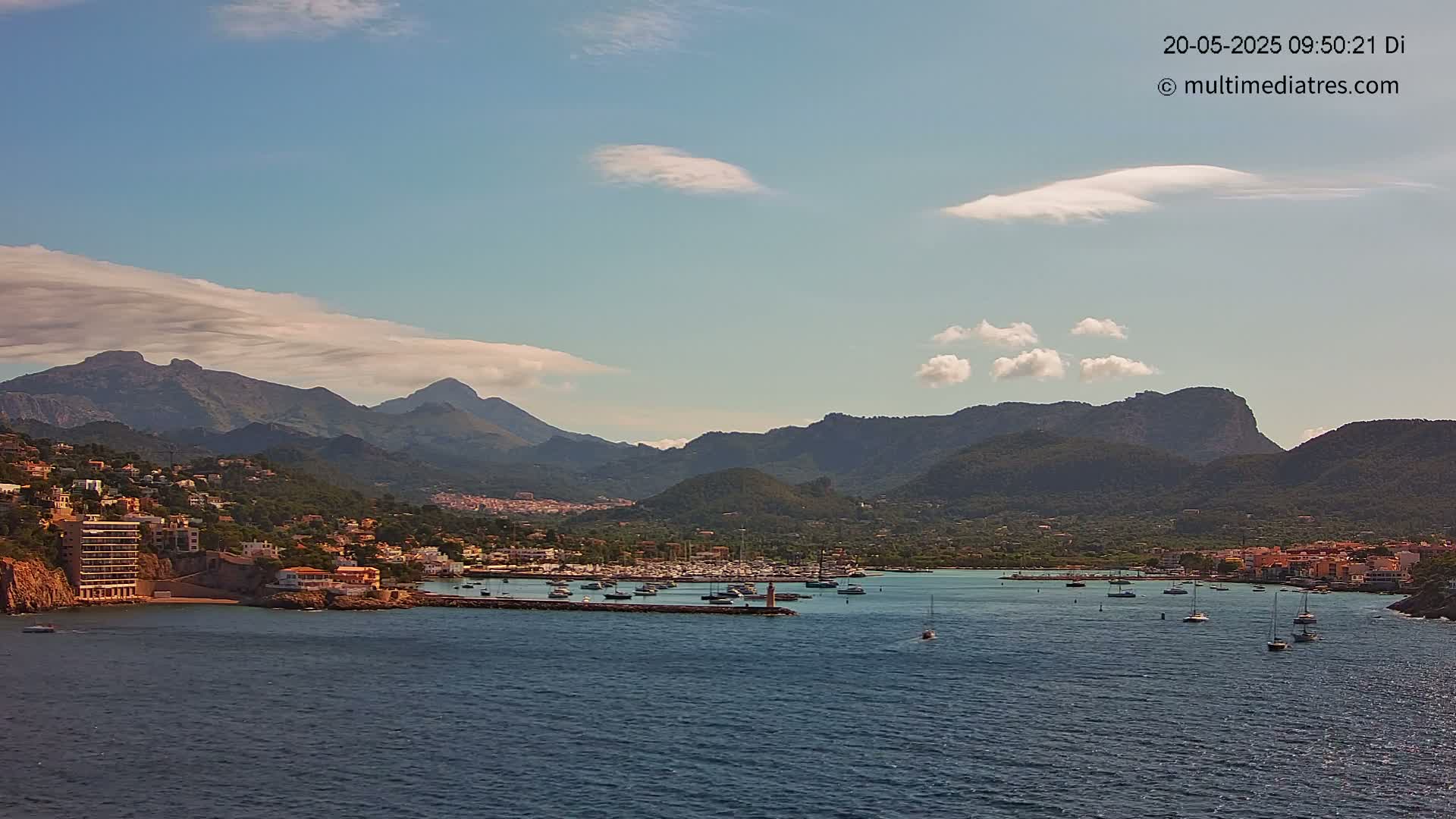 A calm bay with numerous boats is nestled between a hillside town and a mountain range under a partly cloudy sky.