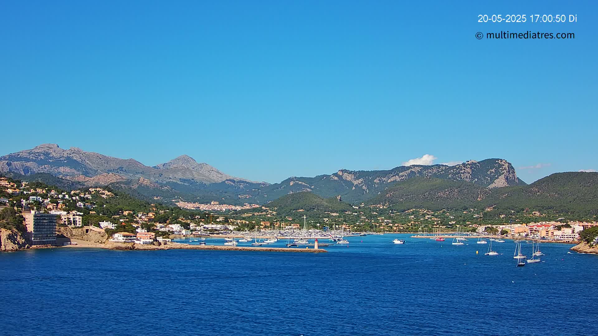 A coastal town nestled against a mountain range under a clear blue sky, with a marina filled with boats in calm waters.