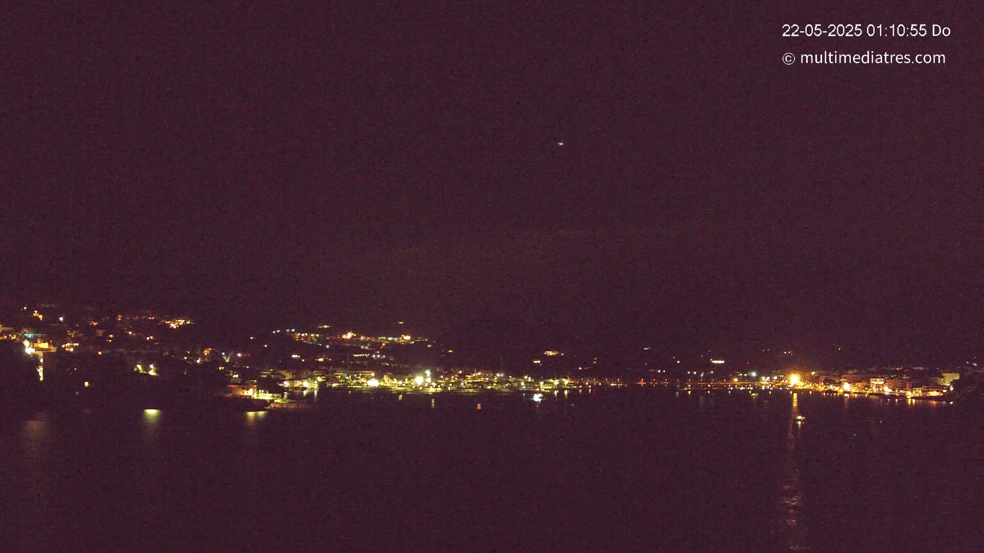 A nighttime long shot shows a dark body of water reflecting the lights of a town nestled on a hillside under a clear, dark sky.