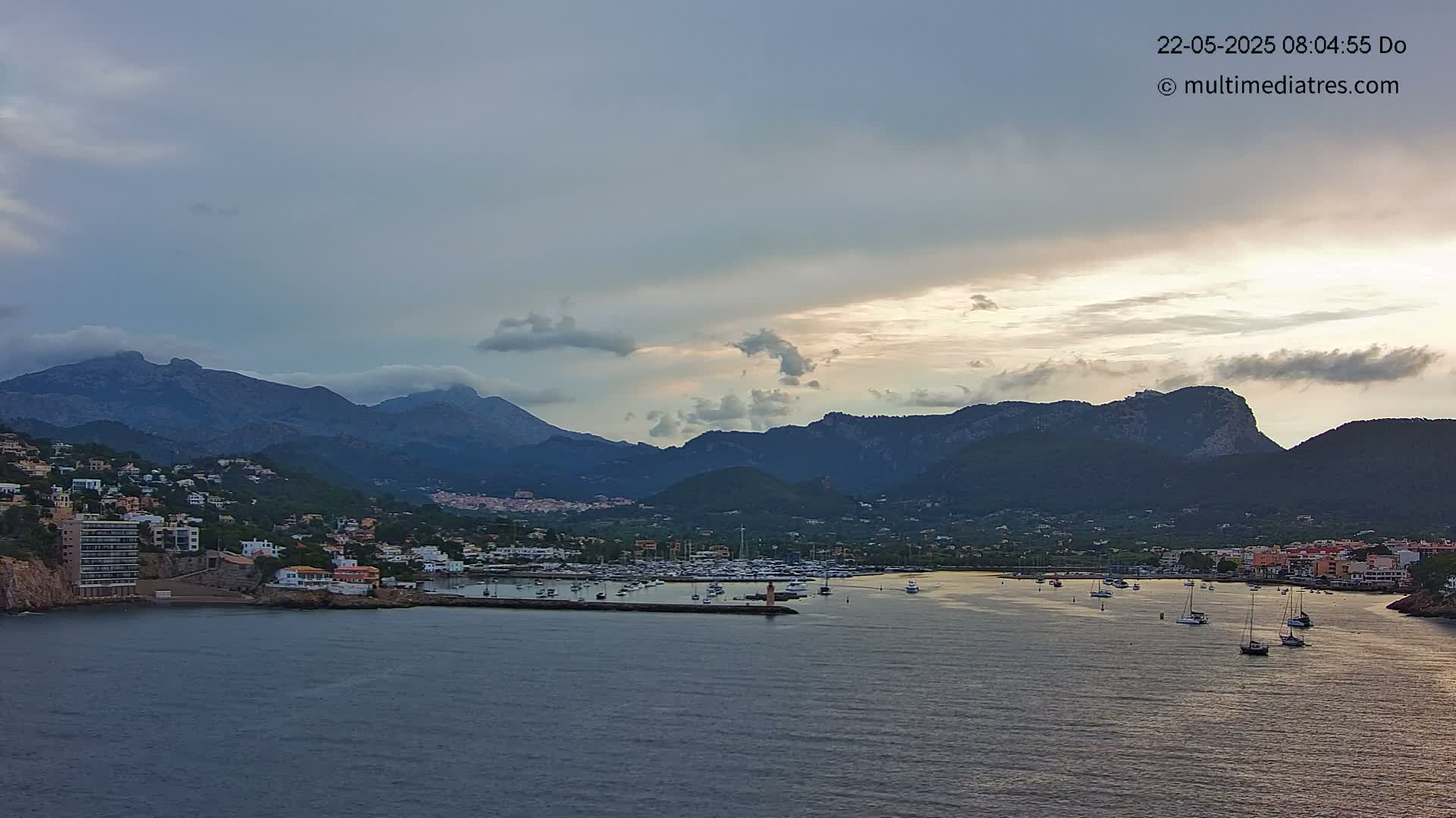 A coastal town nestled between mountains under a partly cloudy sky, with a calm harbor filled with boats.