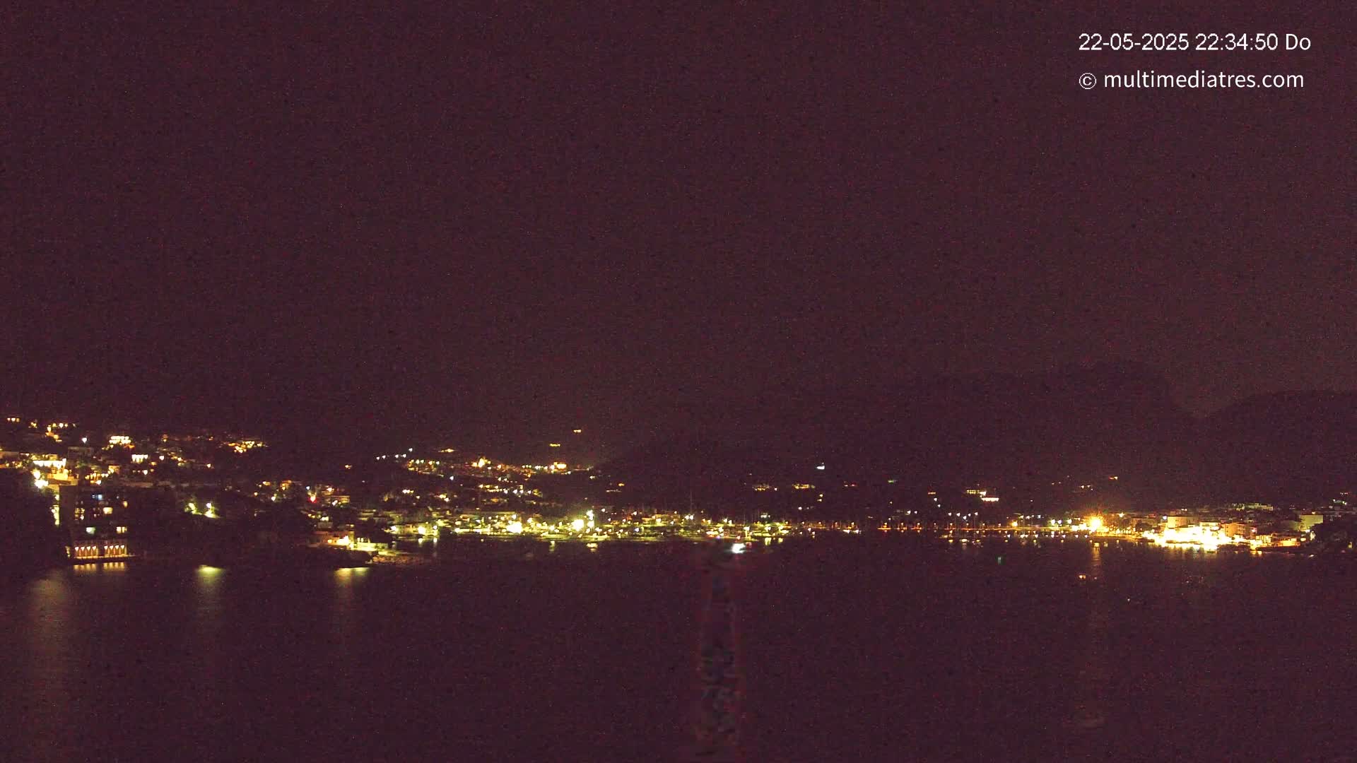 A nighttime, long-range view of a town illuminated by lights along a dark body of water, under a clear, dark sky.