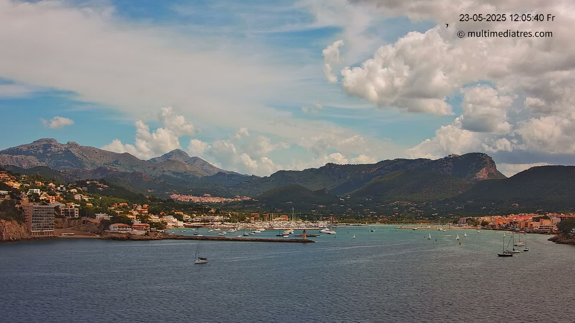 A coastal town nestled at the base of mountains, with a harbor filled with boats, under a partly cloudy sky.