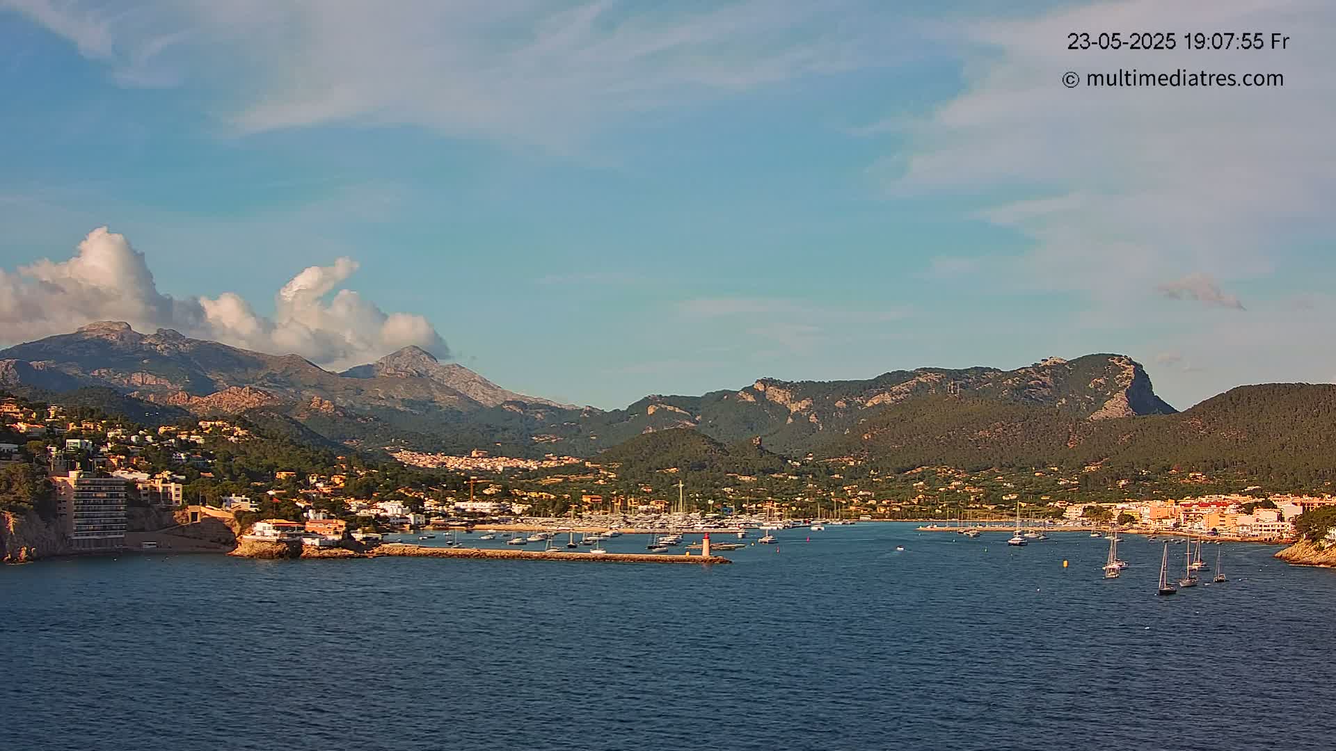 A coastal town nestled between mountains under a mostly clear sky with some cumulus clouds, features a harbor with numerous boats.