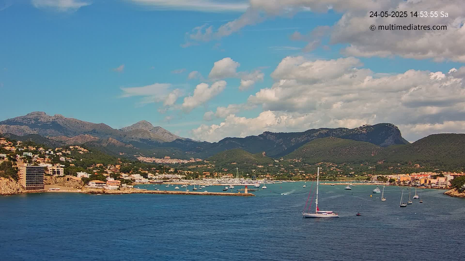 A partly cloudy sky overlooks a harbor filled with sailboats and motorboats nestled in a coastal town, backed by a range of mountains.