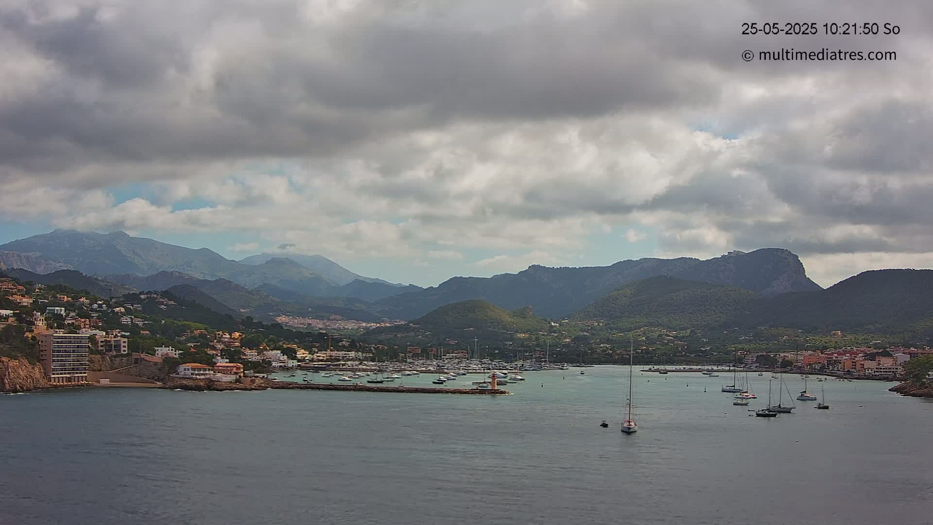 A coastal town nestled among mountains, with a harbor full of sailboats, under a mostly cloudy sky.