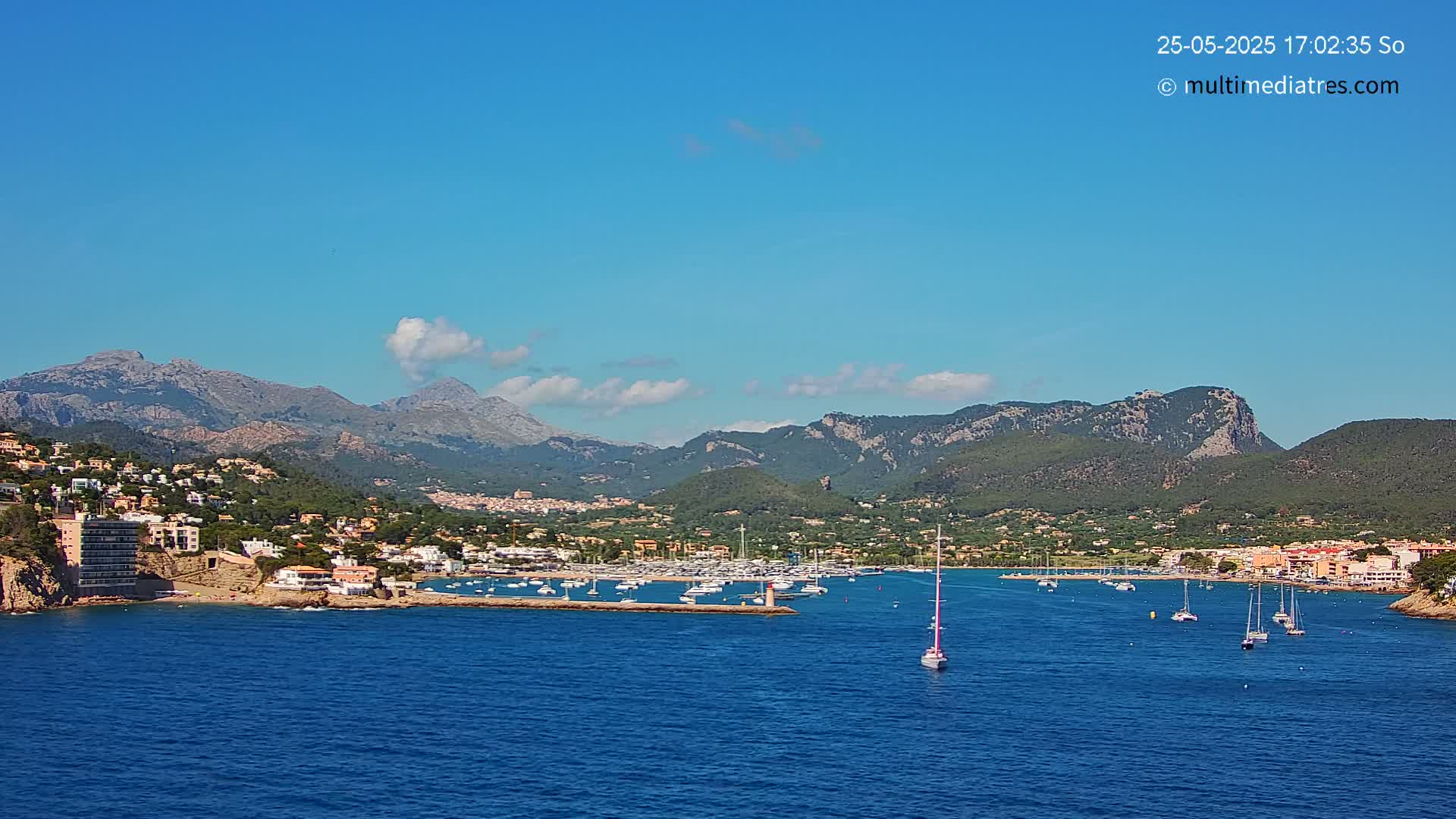 A picturesque coastal town nestled in a mountainous region, with a marina filled with boats and a clear blue sky with a few scattered clouds.