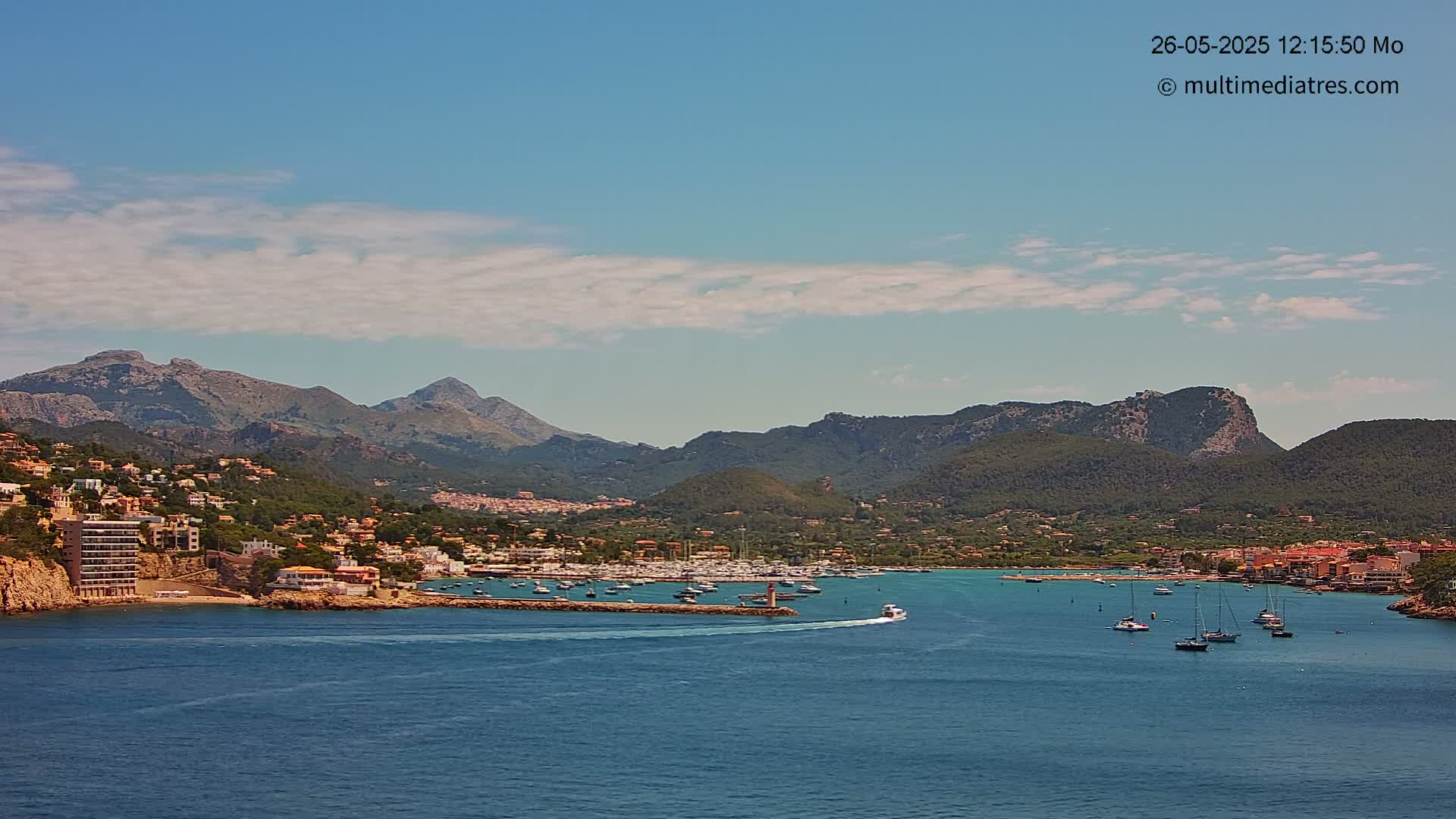 A coastal town nestled against a mountain range under a mostly sunny sky with scattered clouds, features a harbor filled with boats.