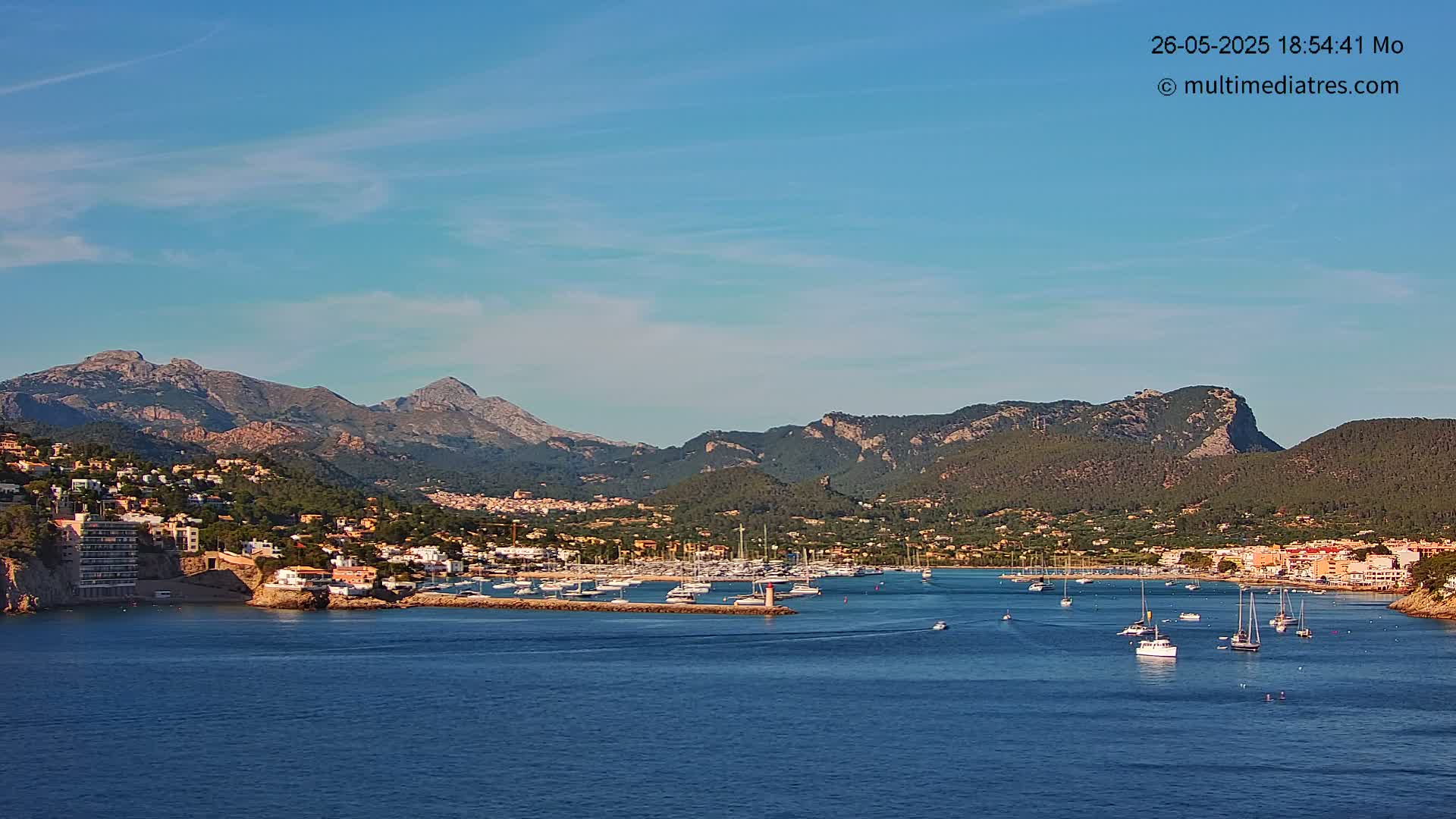 A harbor filled with sailboats is nestled in a coastal town at the foot of a mountain range under a clear blue sky.