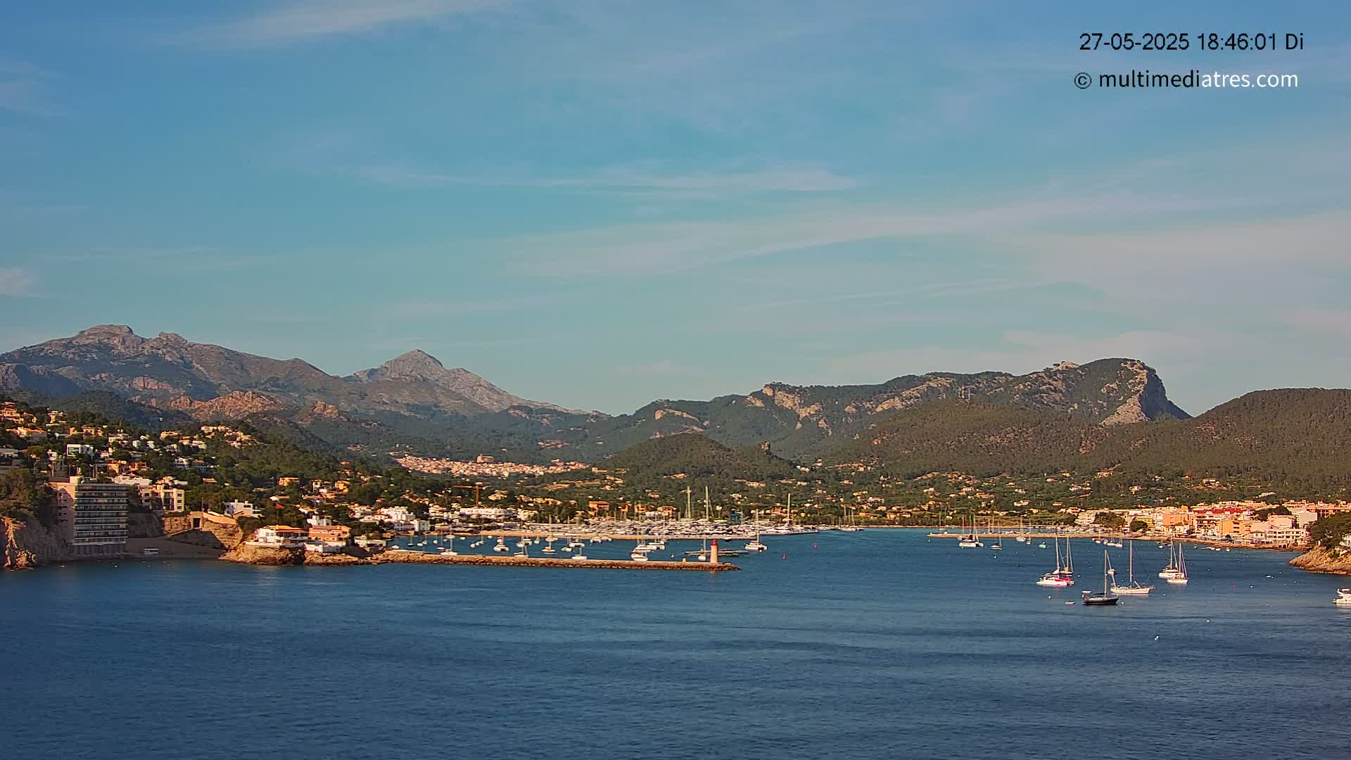 A harbor filled with sailboats sits nestled between a coastal town and a range of mountains under a clear blue sky.