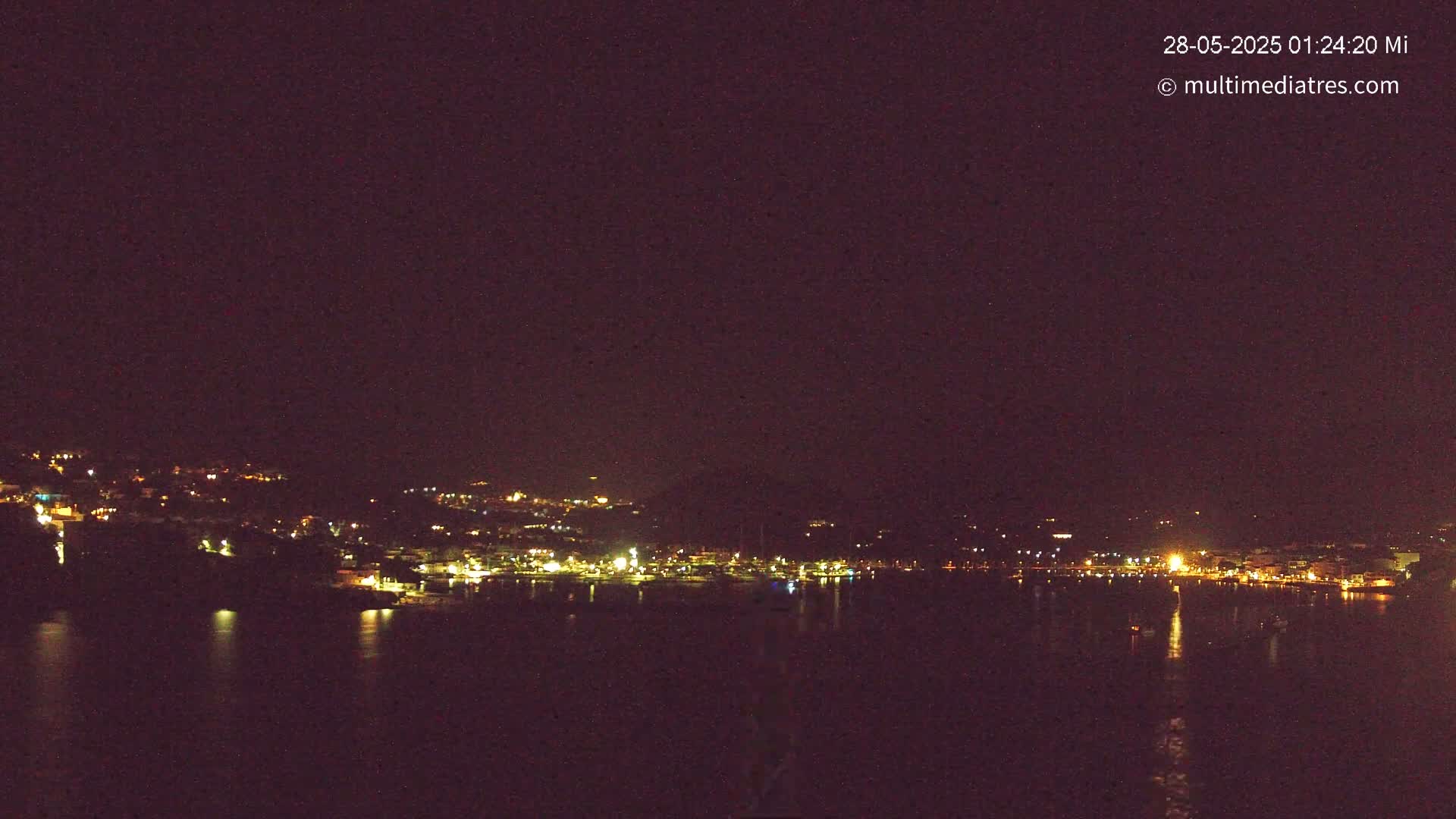 A nighttime view of a coastal town with its lights reflecting on a dark body of water under a clear, dark sky.