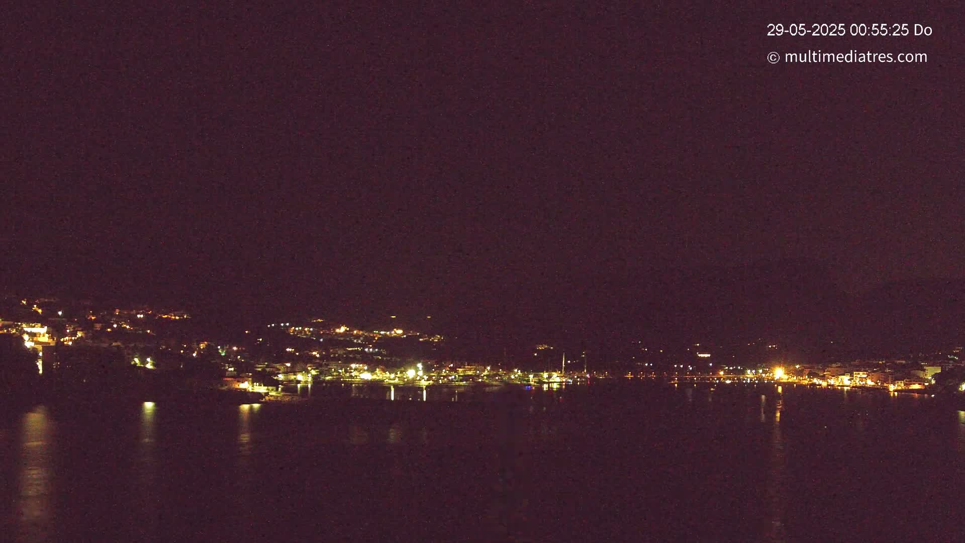 A nighttime view of a town's lights spread across a hillside overlooking a dark body of water under a clear, dark sky.