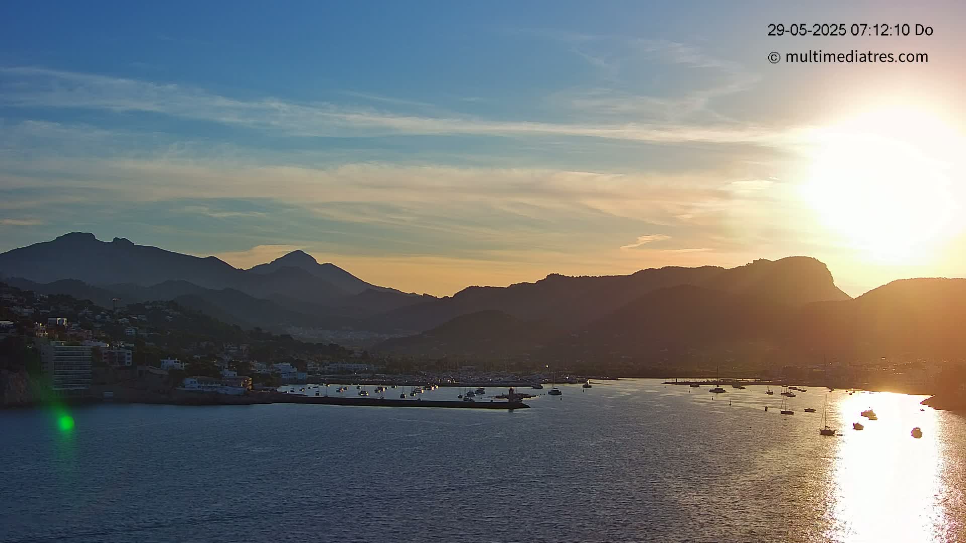 A coastal town nestled between mountains at sunset, with numerous boats in a calm harbor.