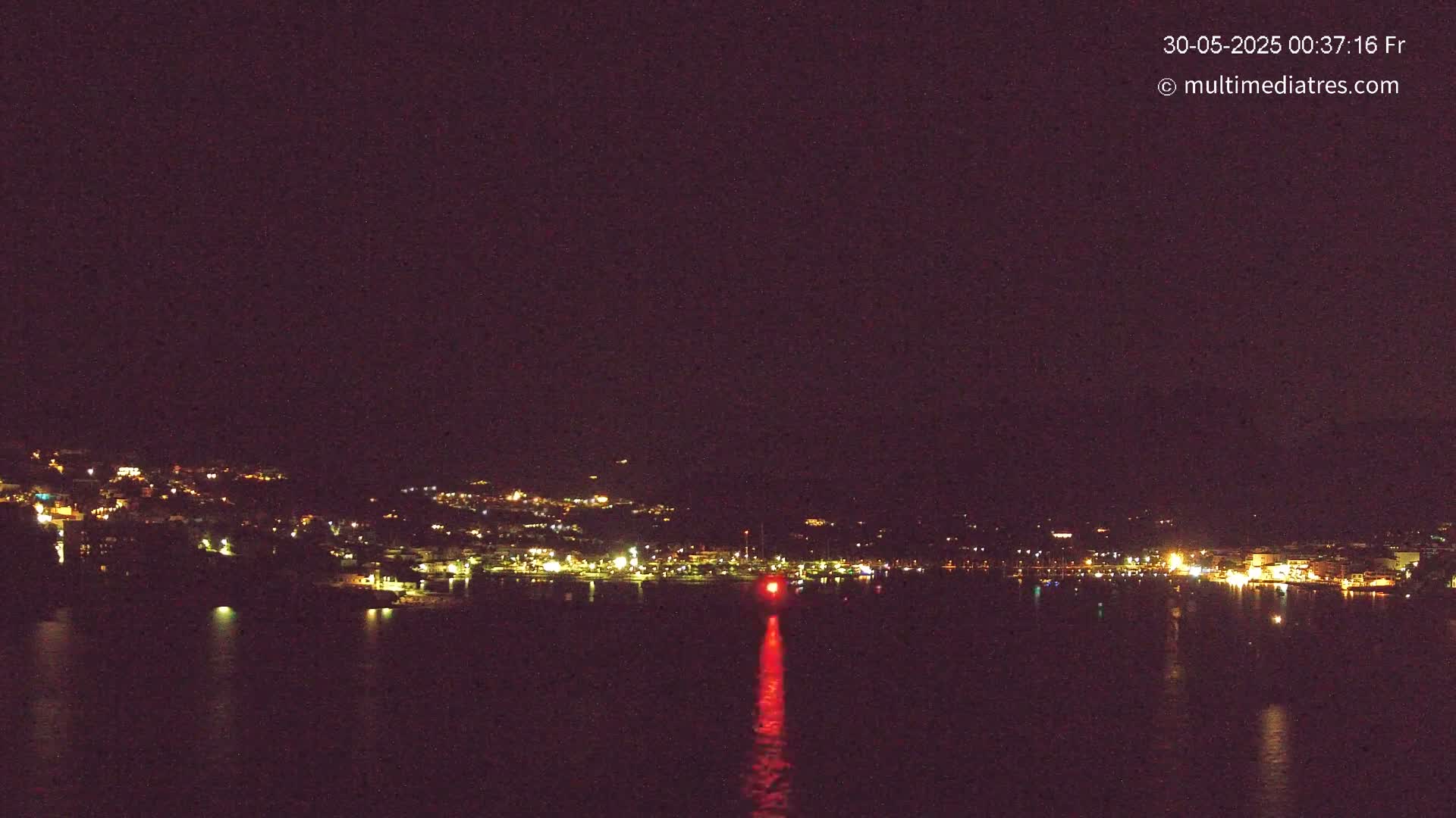 A nighttime view shows a town's lights across a dark body of water, with a single red light reflecting on the water in the foreground.