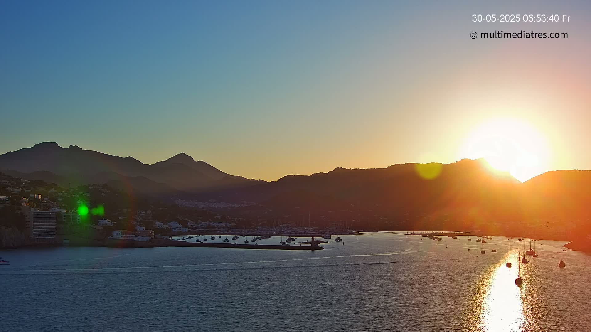 A coastal town nestled between mountains is bathed in the golden light of a setting sun over calm waters dotted with boats.